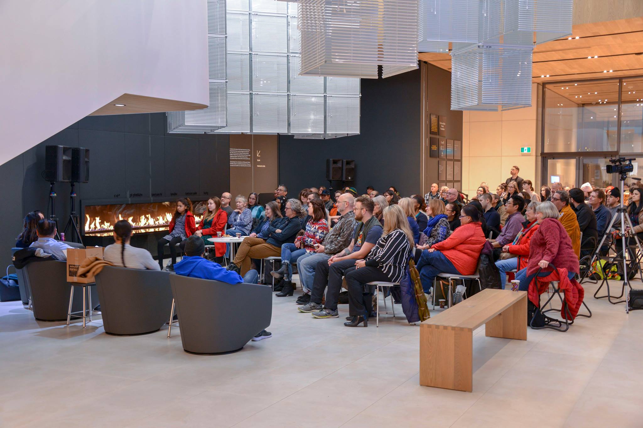 A group of people sit on chairs gathered in Remai Modern's atrium. A fireplace is lit in the background.