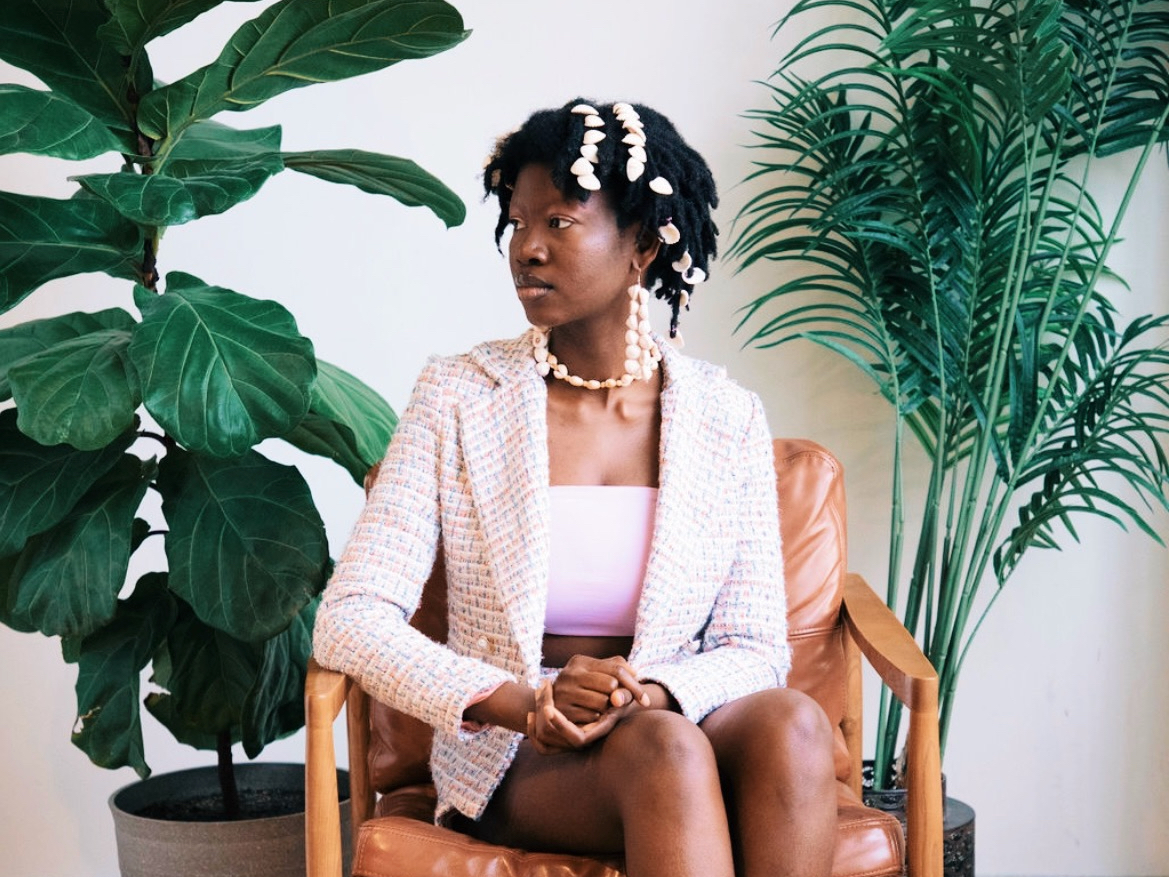 Filmmaker Miley Afolabi sits and looks to the left. Two large plants stand on either side.