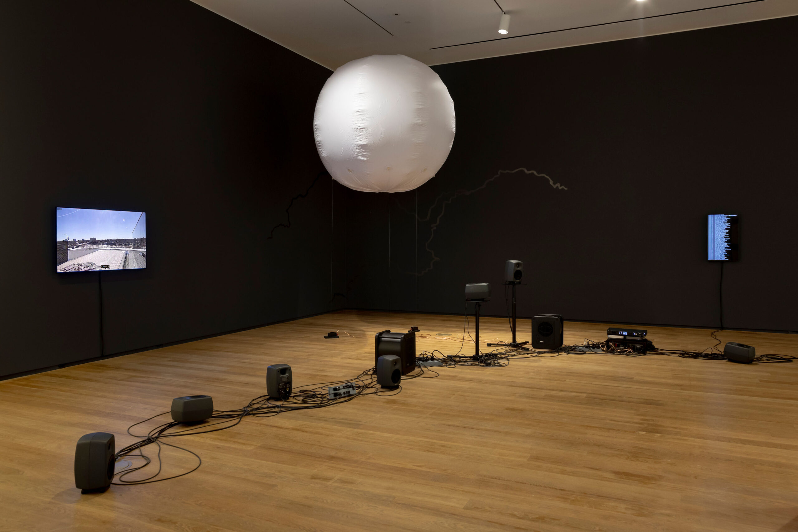 A weather balloon is suspended in the air, surrounded by two screens and an assortment of speakers and cables.