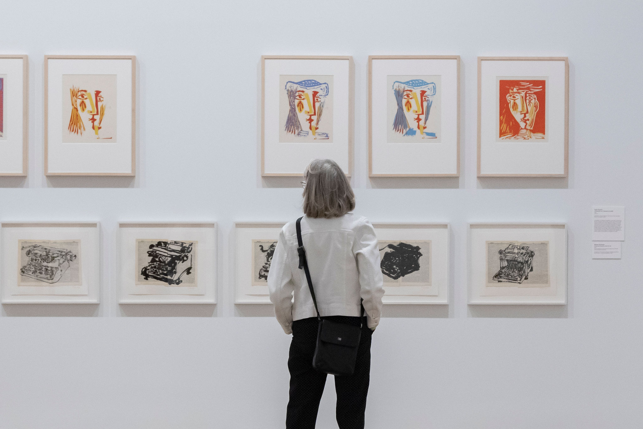A viewer with a grey bob looks at colourful portraits by Pablo Picasso, hung overtop black-and-white prints by William Kentridge.