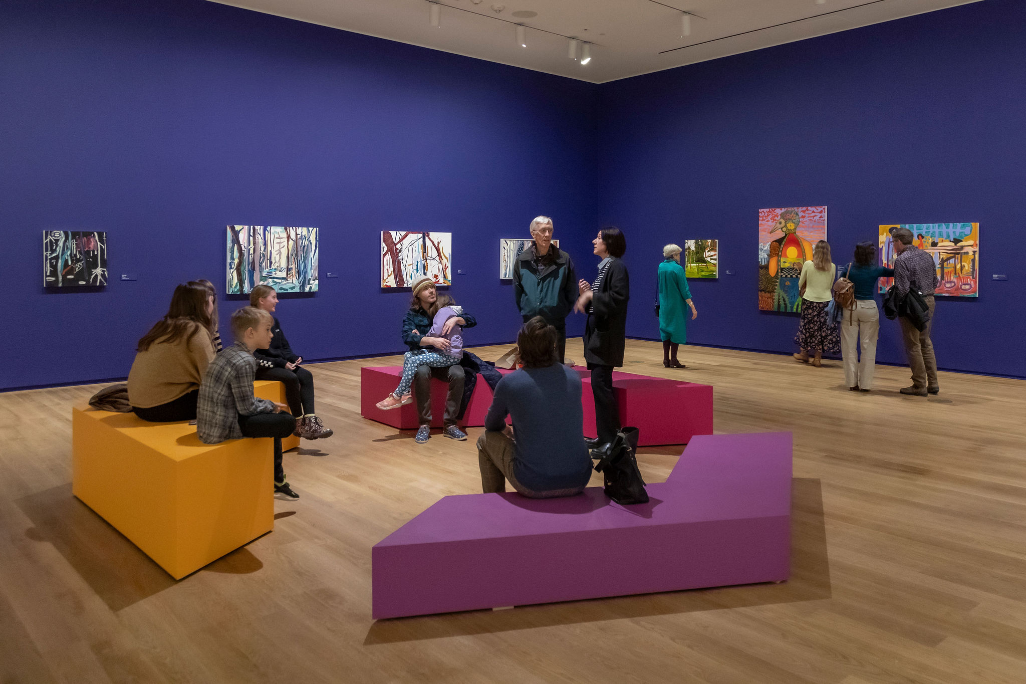 People sit on and stand around three, abstract-shaped benches in yellow, purple and fuchsia. In the background, numerous paintings hang on a wall painted deep purple.