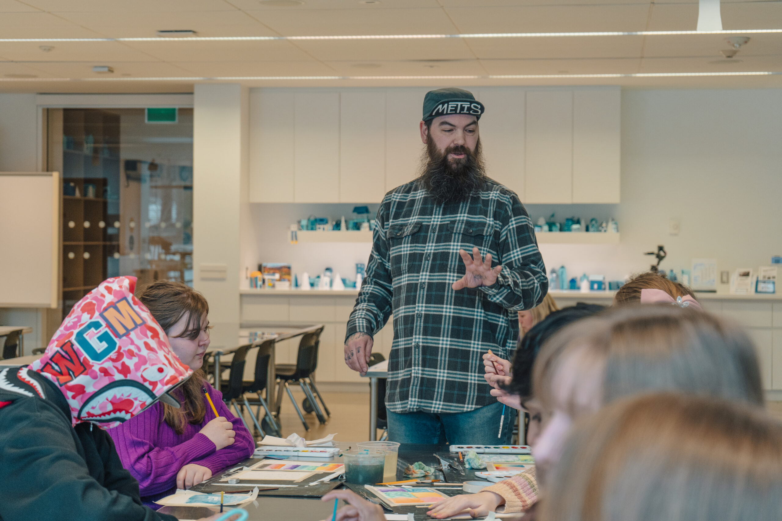 Métis artist Dylan Primeau leads a Youth Art Night workshop at Remai Modern. He stands at the head of a table surrounded by youth participants, gesturing with one hand while speaking. The group is engaged in painting activities, with colourful artwork and supplies spread out on the table. Primeau wears a plaid shirt and a cap that reads “MÉTIS.” The atmosphere is creative and collaborative, set in a bright, modern classroom space.