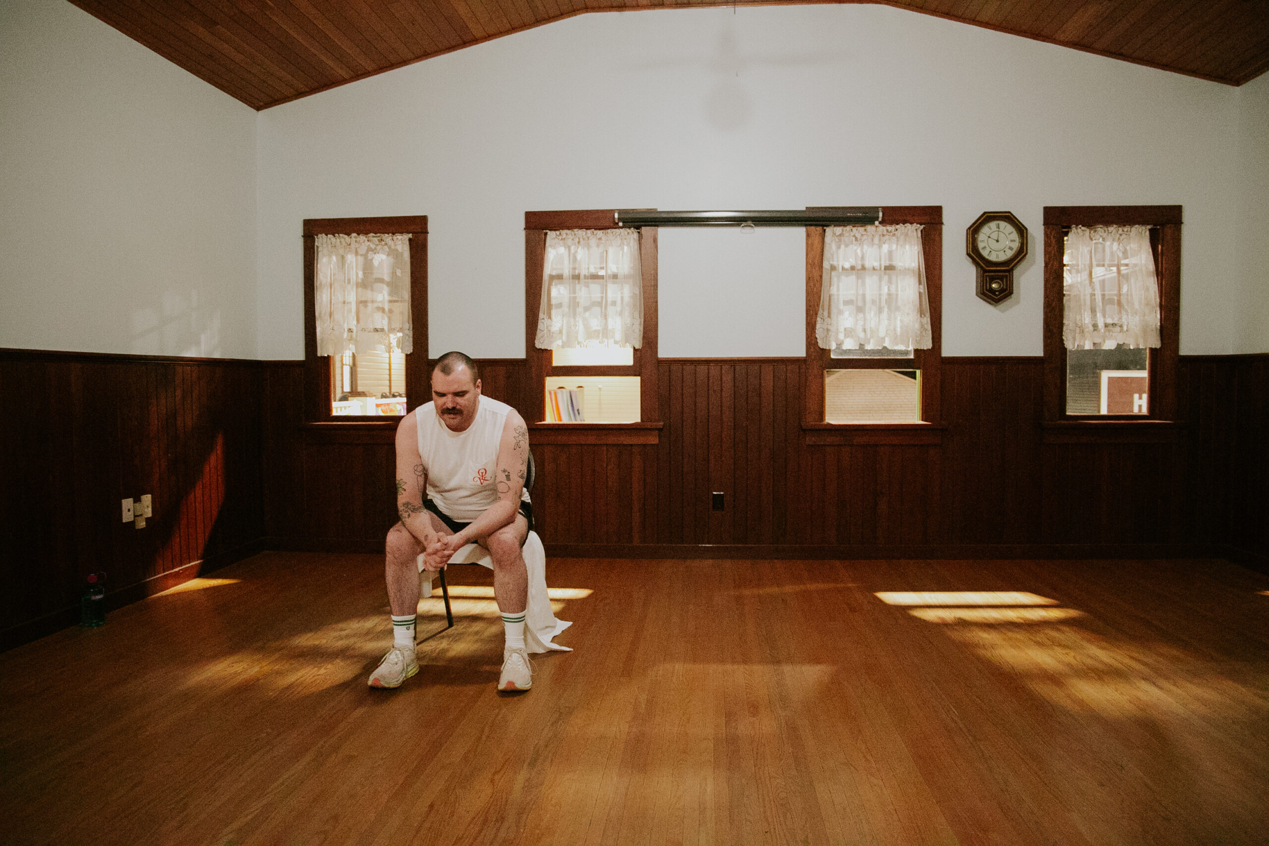 Artist Luke Maddaford sits alone on a chair in the centre of a sparsely furnished room with wood-paneled walls and a vaulted ceiling. Wearing a white tank top, athletic shorts, and sneakers, Maddaford leans forward with his elbows on his knees, his expression contemplative. The soft, warm lighting highlights the vintage feel of the space, with lace curtains on the windows and a traditional wall clock above. The scene conveys a sense of stillness and introspection.