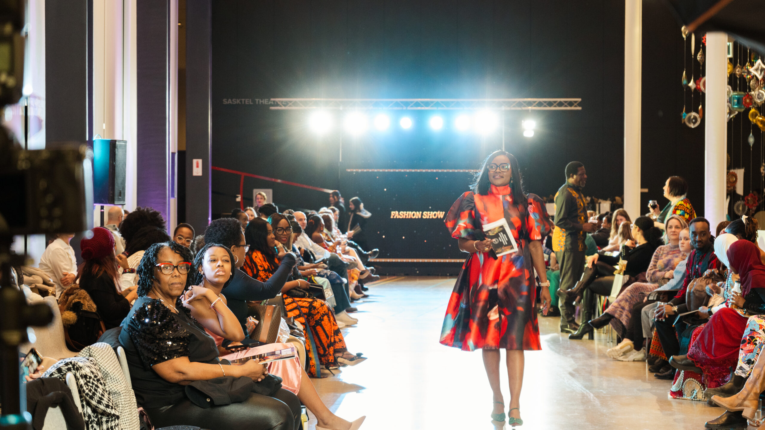 A confident woman in a bold red and black patterned dress walks down a brightly lit runway holding a booklet. She is surrounded by a diverse audience seated on both sides, many dressed in vibrant attire. Spotlights shine behind her, and a black backdrop reads “FASHION SHOW” in glowing letters. The atmosphere is lively and celebratory.