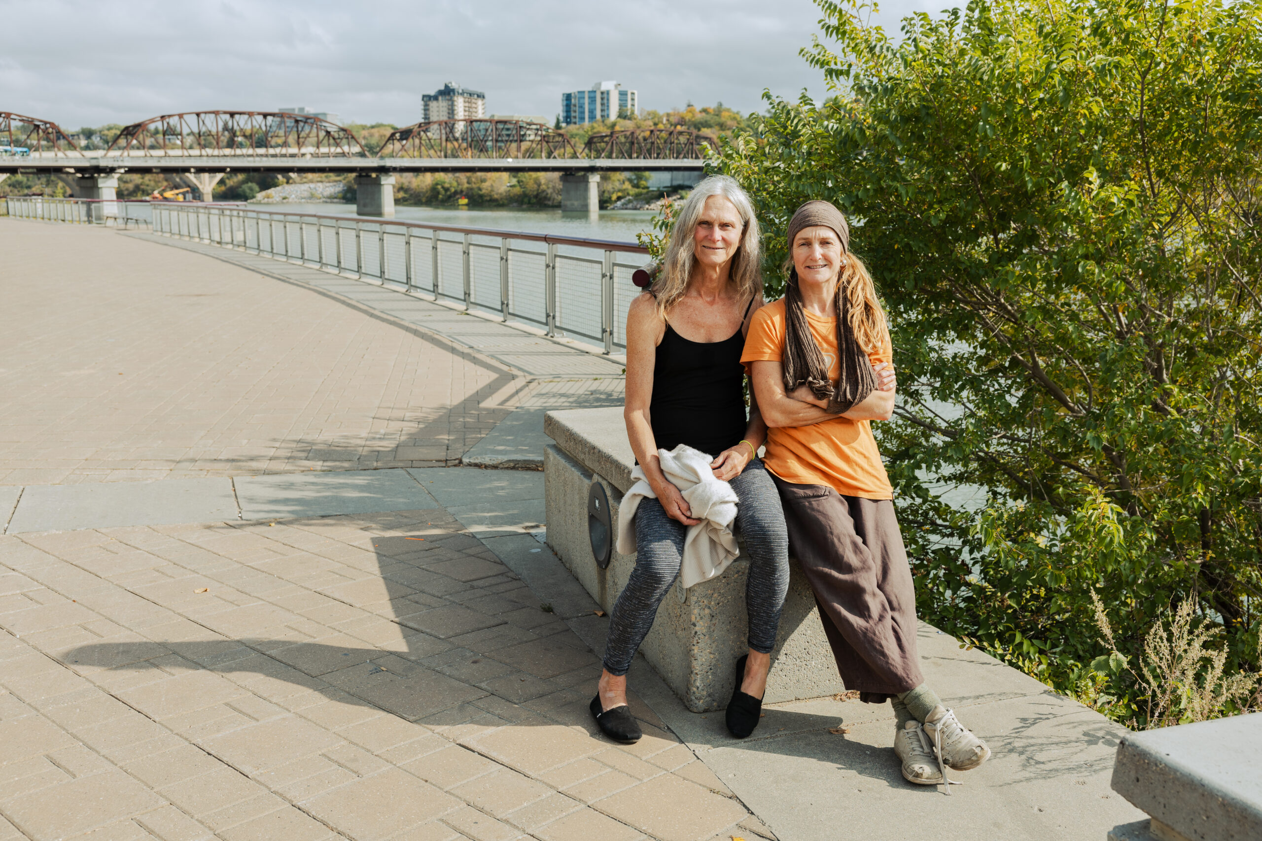 Artists Miki Mappin (left) and Kyle Syverson (right) of KSAMB Dance Company sit side by side on a riverside promenade bench in Saskatoon. Mappin wears a black tank top and grey leggings, holding a light sweater, while Syverson wears an orange shirt, brown pants, and a scarf wrapped around their head and shoulders. Behind them, the South Saskatchewan River and a historic traffic bridge stretch across the frame under a lightly overcast sky.