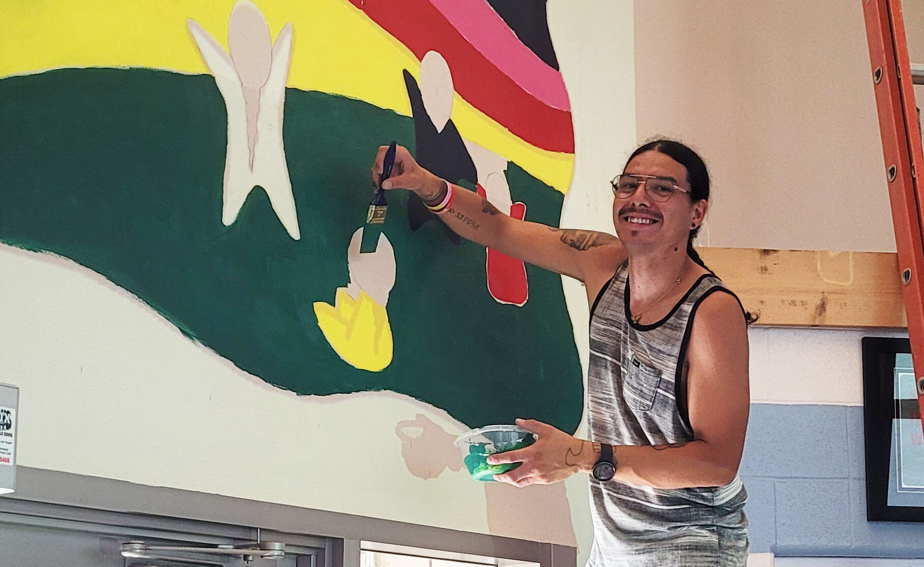 Artist James Desjarlais smiles while painting a colourful mural on a high interior wall. Standing on a ladder in a tank top and shorts, he holds a paintbrush in one hand and a container of green paint in the other. The mural features abstract human figures in front of vibrant horizontal bands of yellow, red, and black, with areas of green and pink still in progress. Natural light streams through the nearby glass door, illuminating his work.
