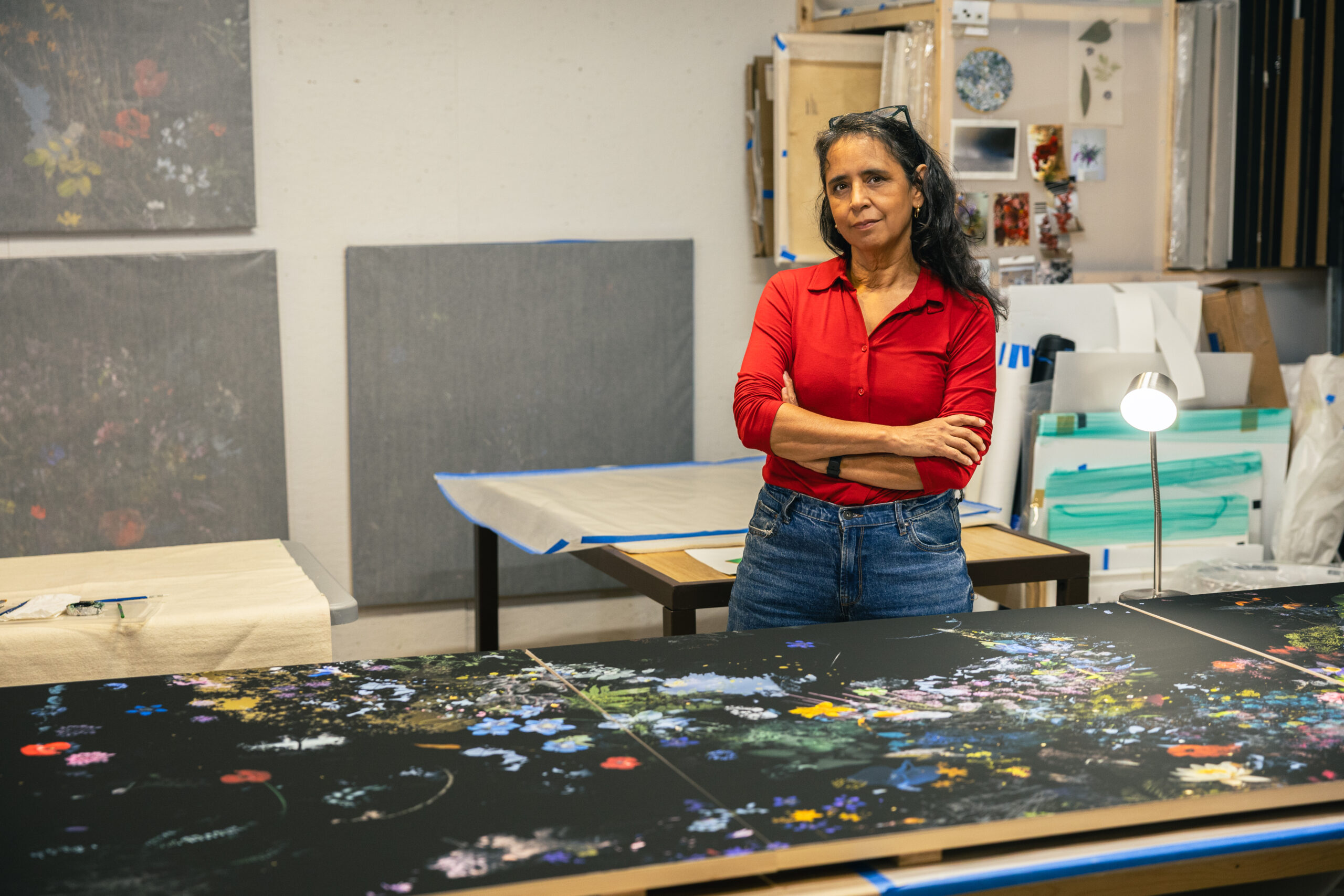 Gabriela García-Luna stands in her art studio with arms crossed, wearing a red shirt and jeans. Behind her are large-scale canvases featuring dark floral compositions, with tools and materials neatly arranged around the space.