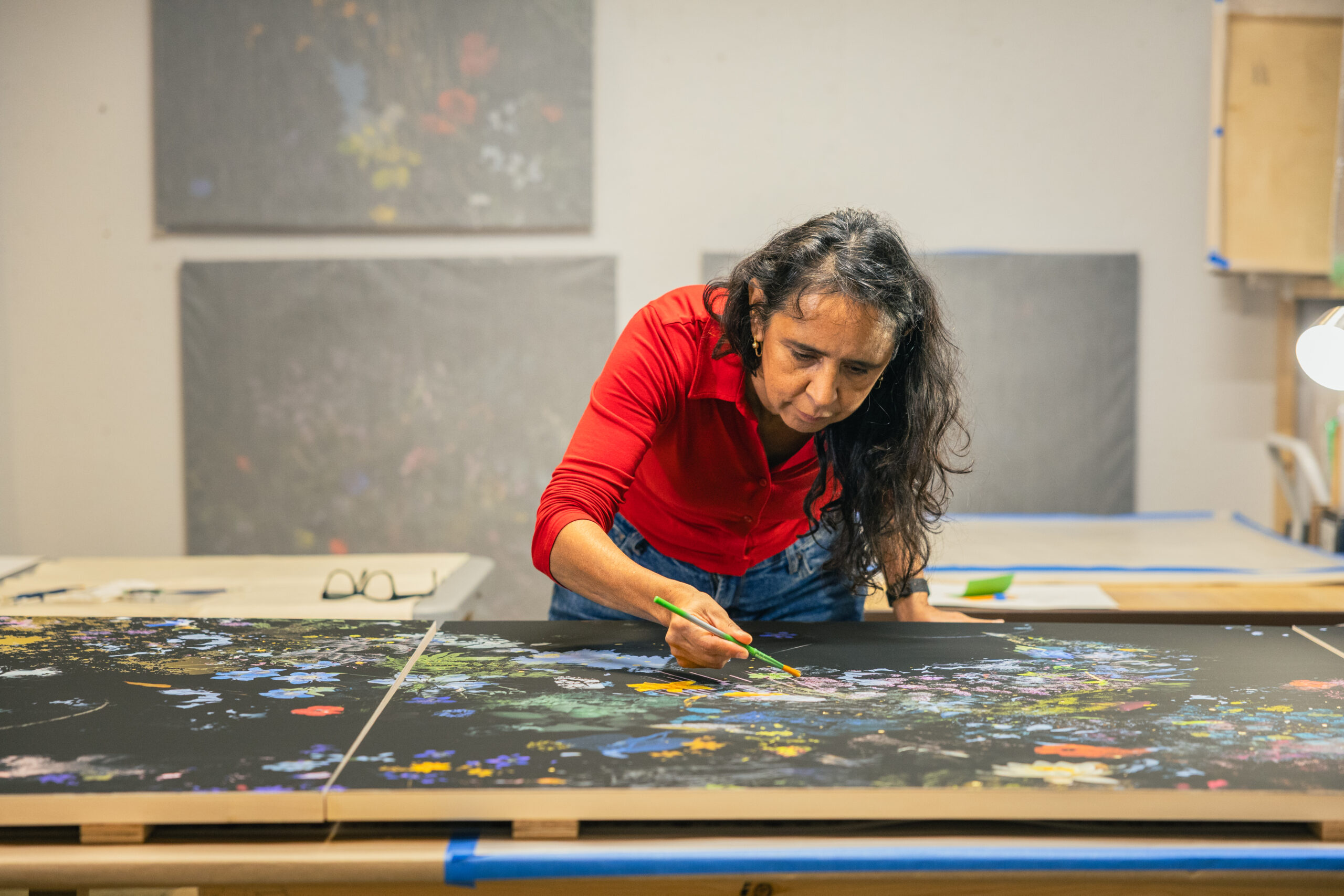 García-Luna concentrates on adding fine details to a vivid, flower-filled painting spread across two black panels. Her bright red shirt contrasts with the intricate colours of the artwork.