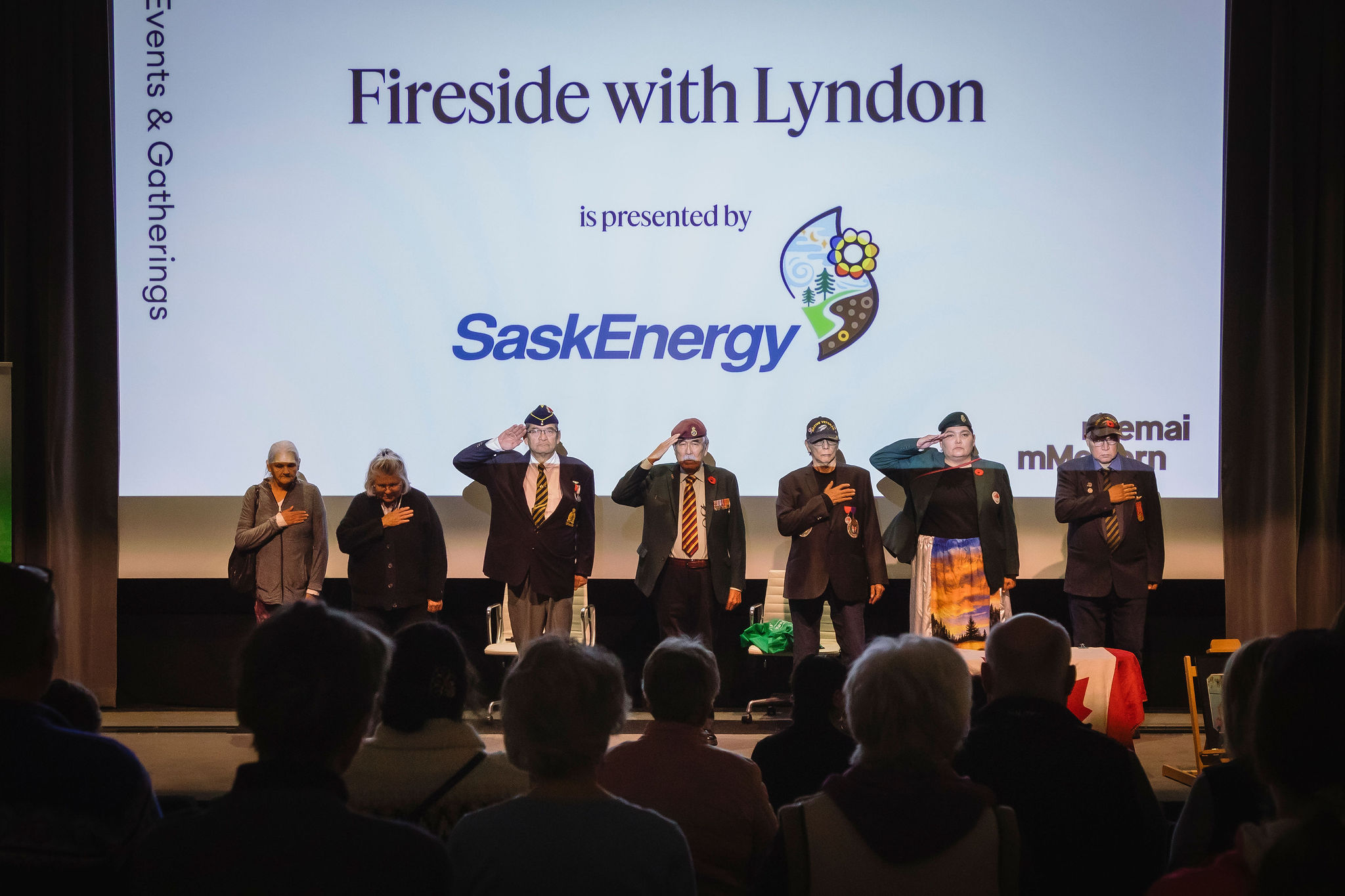 A group of veterans stands on a theatre stage facing an audience, some saluting and others with hands over their hearts.