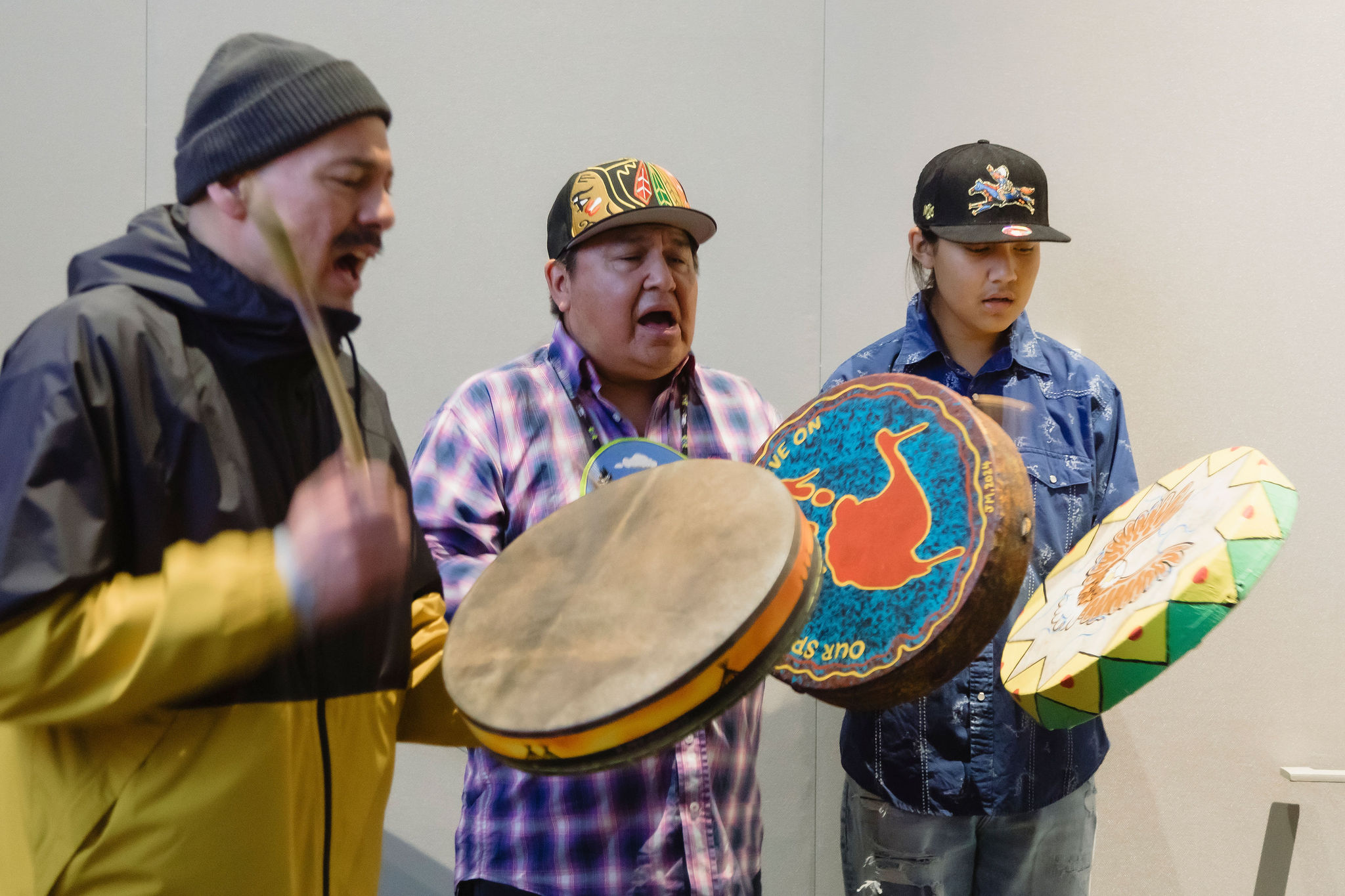 Three people stand indoors, singing and playing hand drums. They wear casual clothing and caps, and each holds a round frame drum decorated with colorful designs.