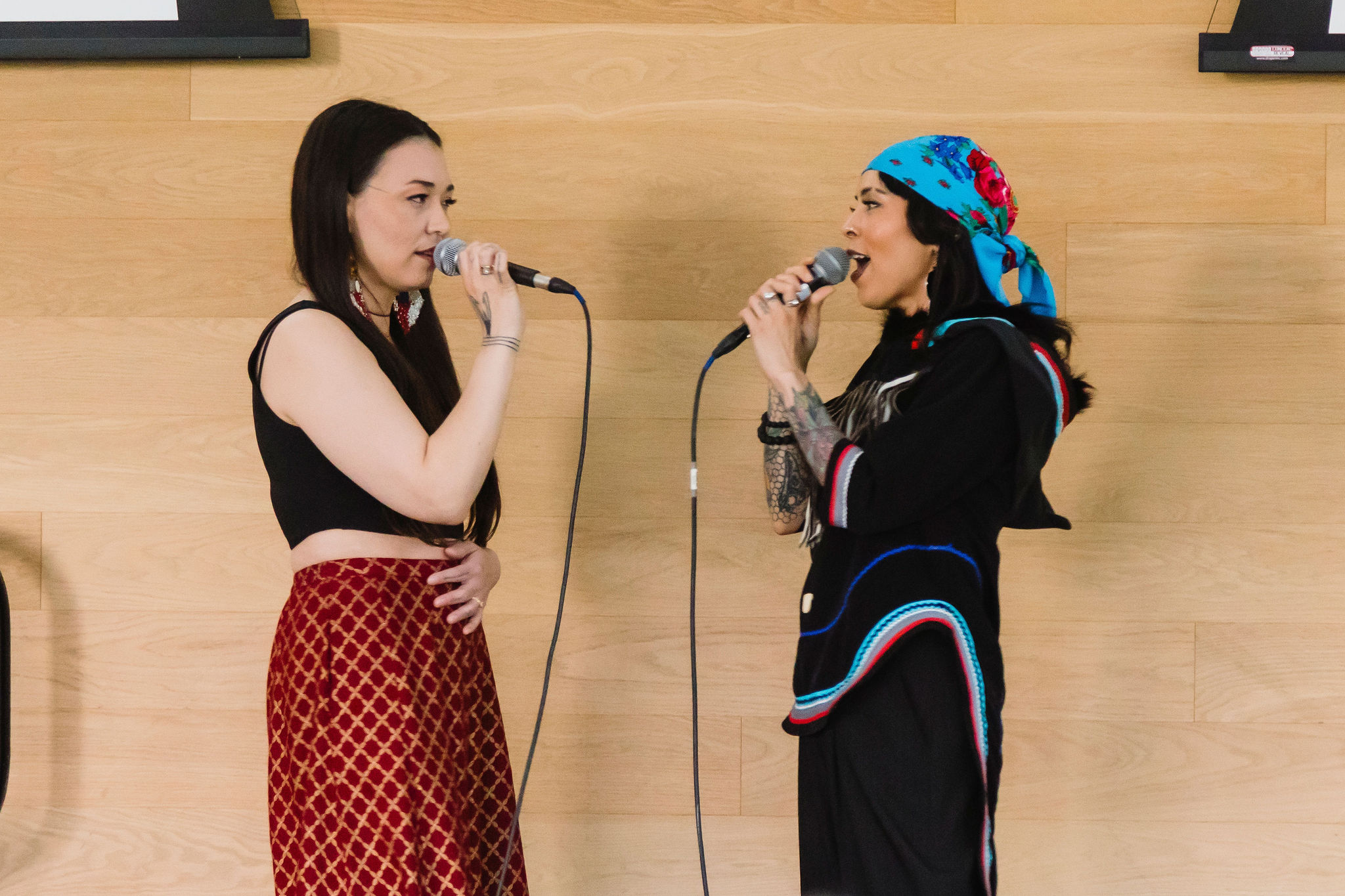 Two performers stand facing each other indoors, each singing into a handheld microphone. One wears a black sleeveless top with a red patterned skirt, while the other wears a black outfit with colourful trim and a blue floral headscarf.