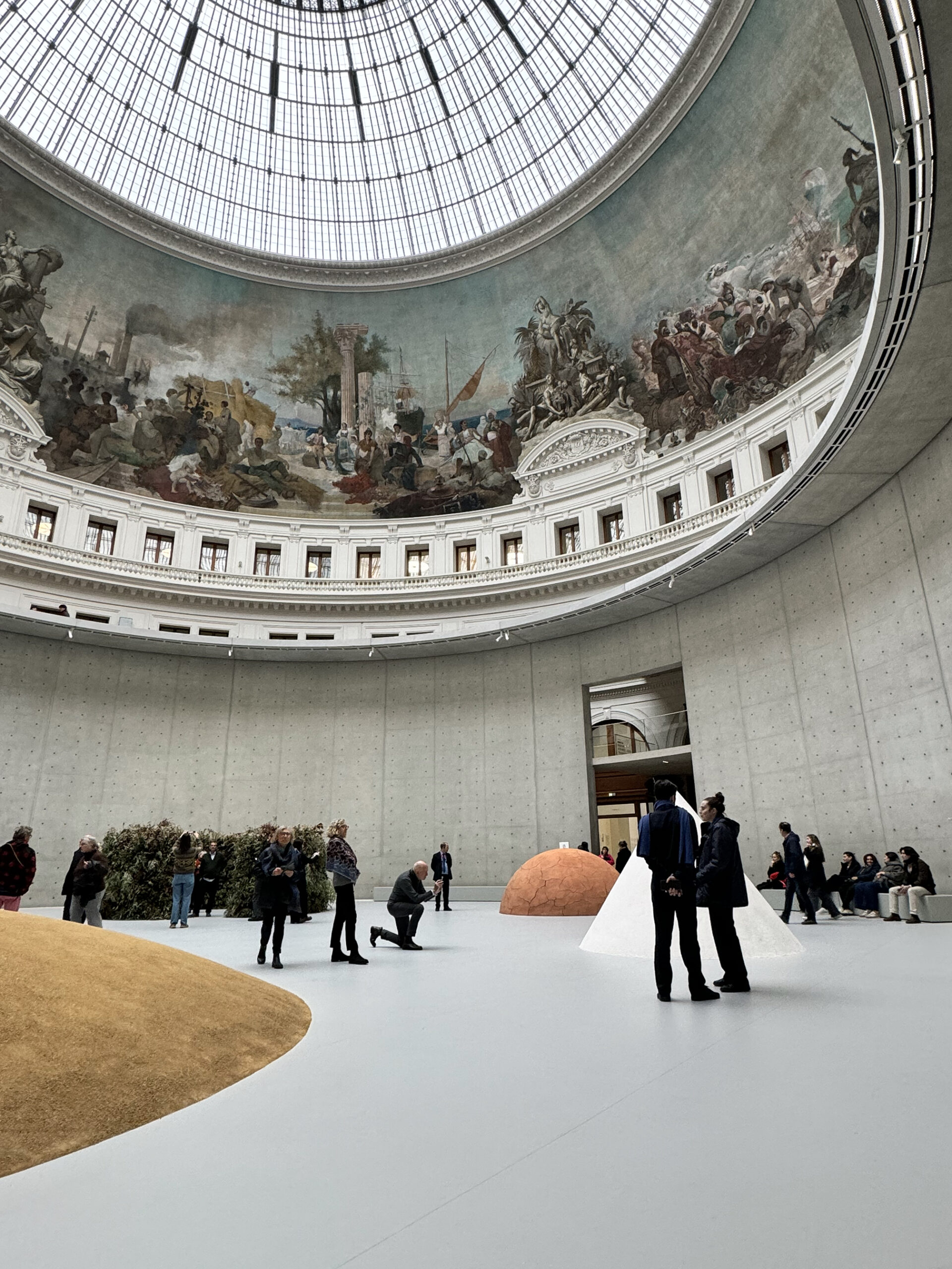 Visitors explore a vast circular gallery beneath a glass-domed ceiling, surrounded by a panoramic mural. Minimal sculptural installations in earth tones sit across the open floor, while people gather, observe, and converse within the monumental museum space.