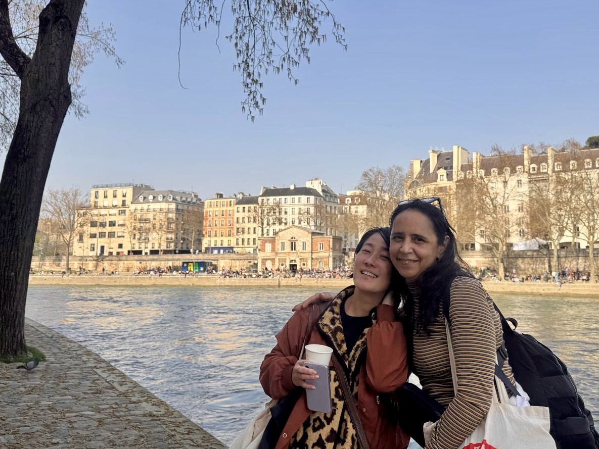 Two people embrace along the Seine on a sunny afternoon, smiling beside the river with Parisian buildings and bare spring trees in the background. One holds a coffee cup while both carry bags, capturing a warm, candid moment during a walk through the city.