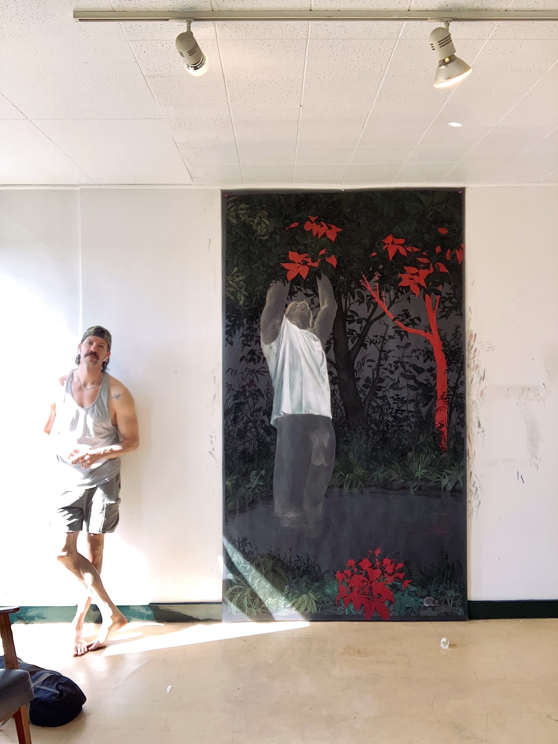 An artist stands barefoot in a sunlit studio beside a large vertical painting mounted on a white wall. The painting depicts a figure submerged waist-deep in dark water, reaching upward into dense foliage illuminated by vivid red leaves and branches. Bright afternoon light floods the room from the left, casting strong highlights and shadows across the floor and the artist’s gray tank top and shorts.
