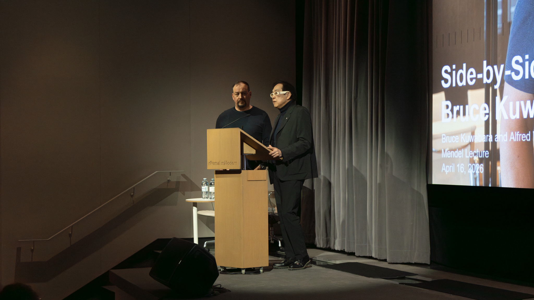 Bruce Kuwabara and Alfred Waugh speaking at the podium during the Mendel International Lecture at Remai Modern. The two architects stand on stage beside a projected title slide for their presentation while addressing the audience in the theatre.