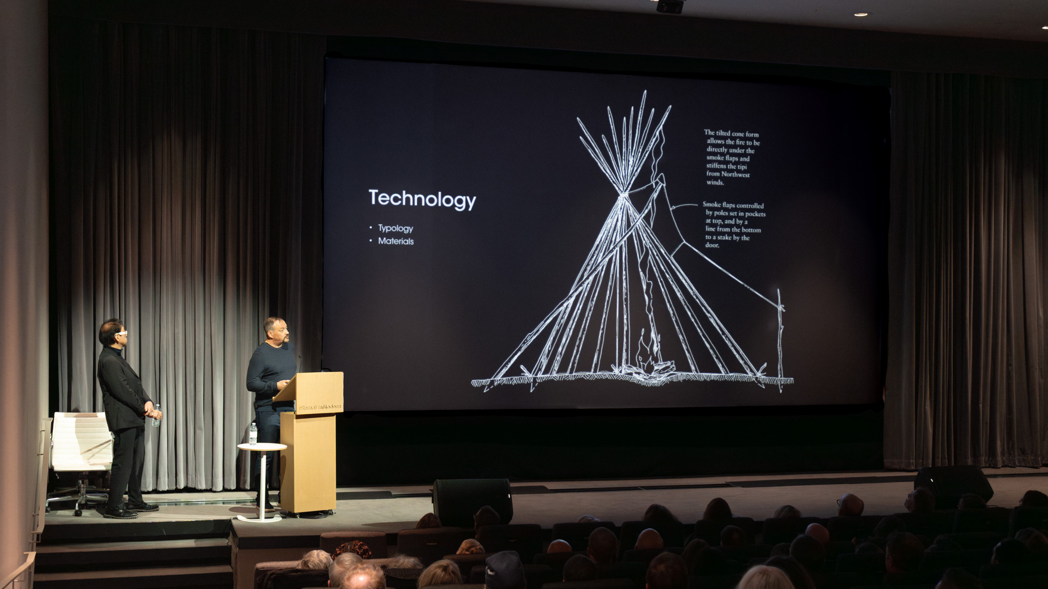 Bruce Kuwabara and Alfred Waugh presenting during the Mendel International Lecture at Remai Modern. A large projection screen displays a slide titled “Technology” alongside a diagram of a tipi structure, while the two architects stand on stage addressing the audience.