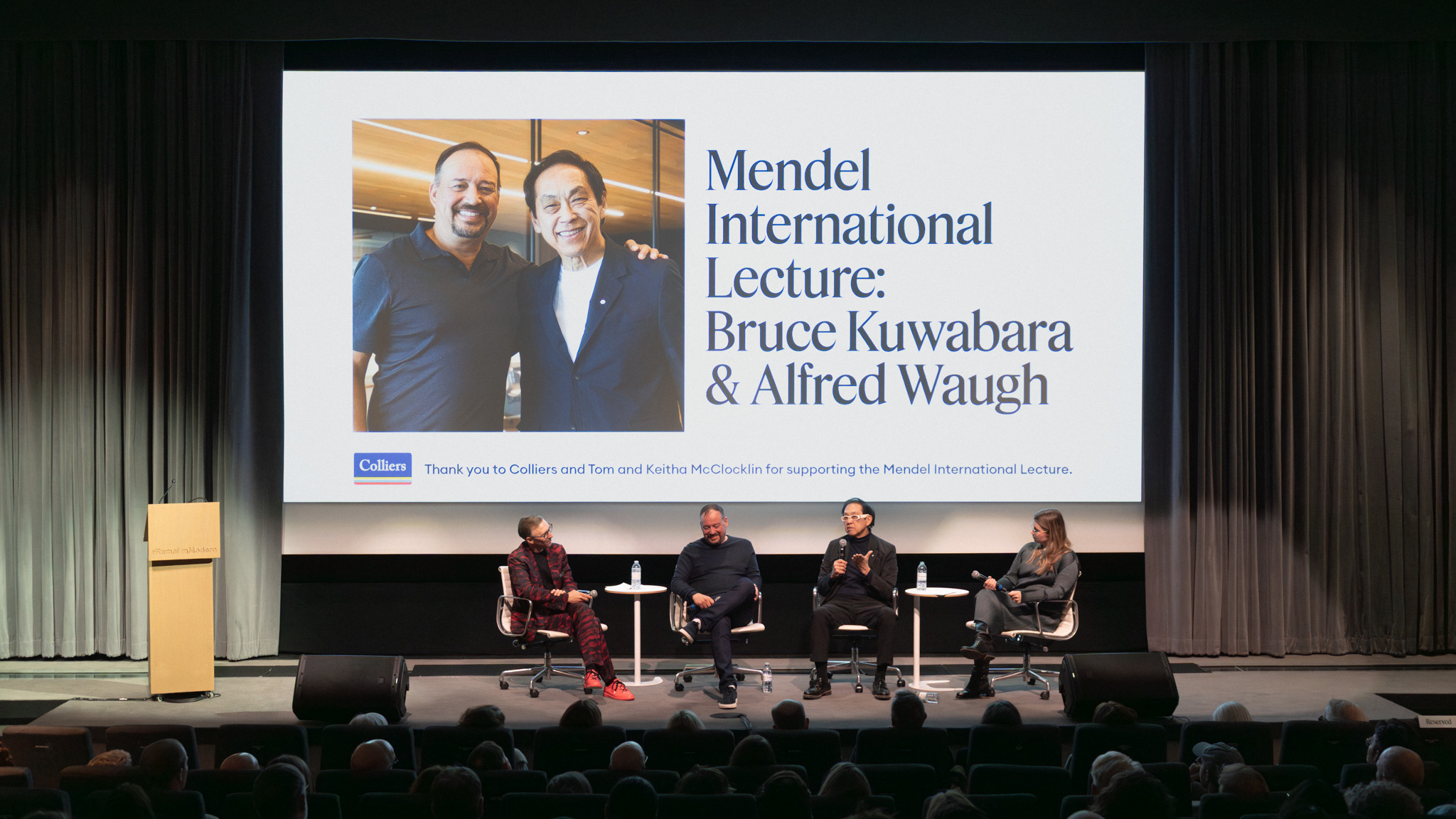 Panel discussion at the Mendel International Lecture featuring Bruce Kuwabara and Alfred Waugh on stage at Remai Modern. A large screen behind the speakers displays event branding and a photo of the two architects, while four panelists sit before an audience in the theatre.