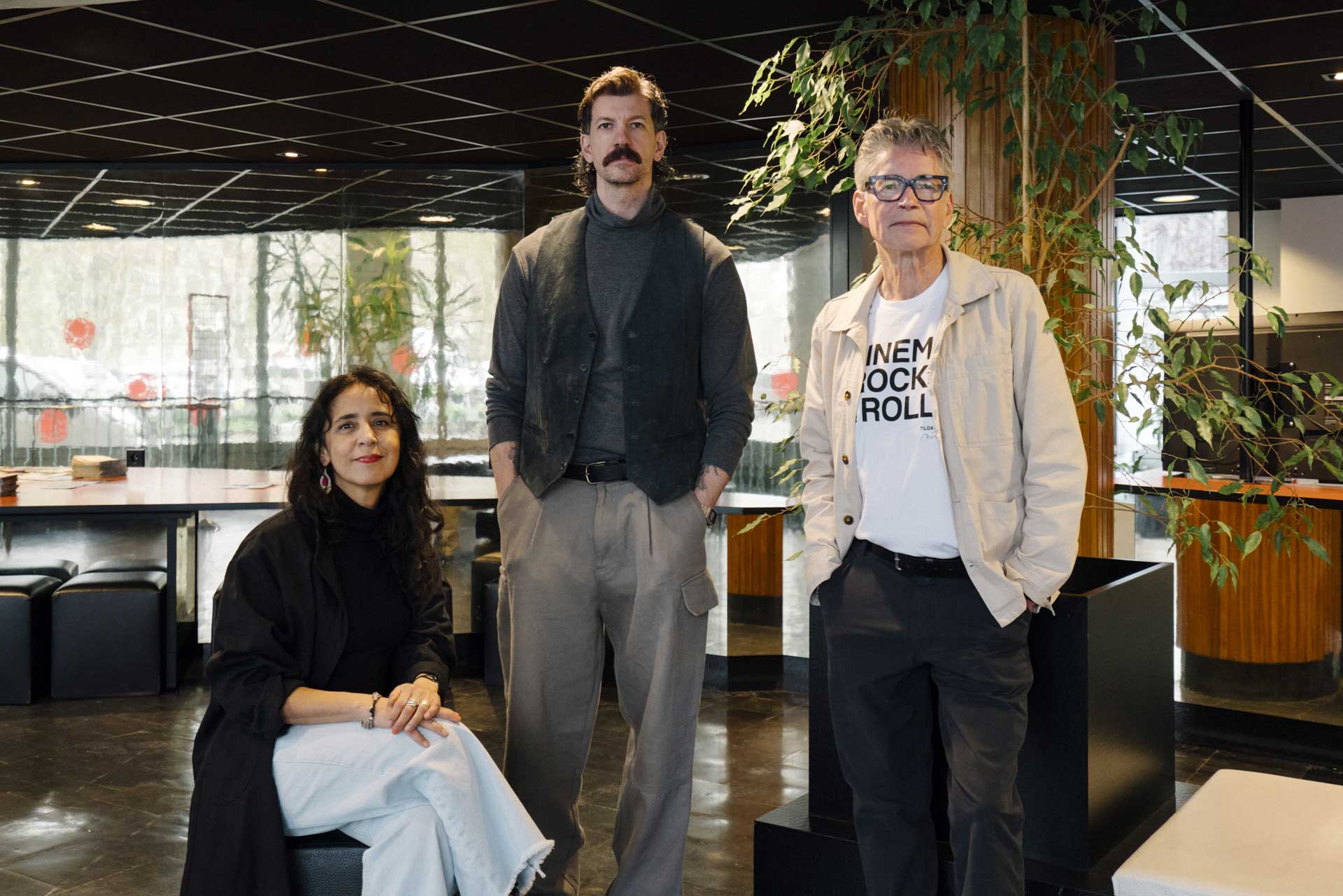 Three people pose together in a stylish modern interior with dark tiled ceilings, reflective walls, and large indoor plants. One woman is seated in the foreground wearing a black top and white pants, while two men stand behind her. The man in the center has a mustache and wears a gray turtleneck with a dark vest and loose gray trousers. The man on the right wears glasses, a light jacket, and a graphic T-shirt.
