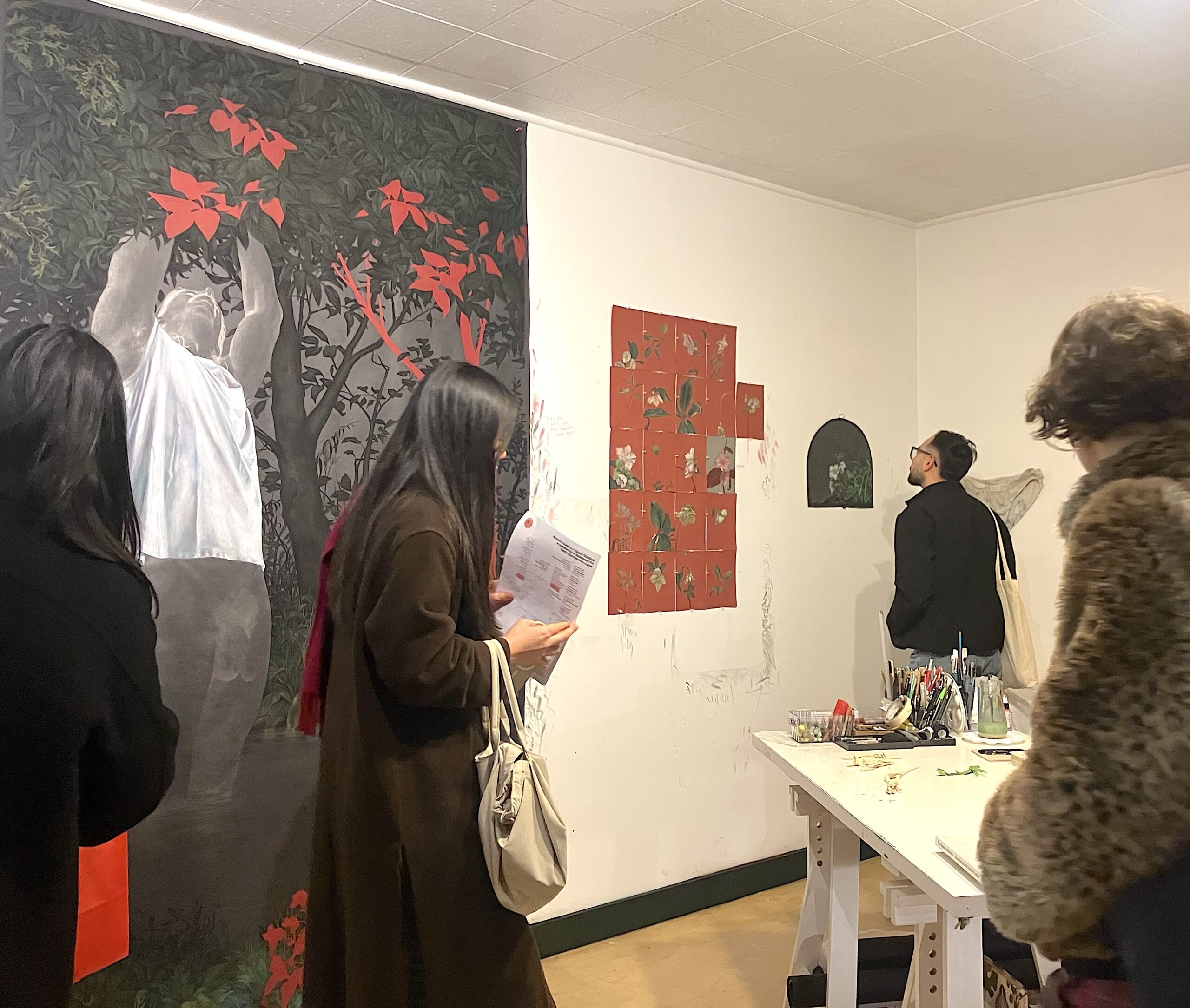 Visitors explore an artist’s studio during what appears to be an open studio or gallery event. A large, striking painting dominates the left wall, showing a figure standing in dark water beneath dense foliage with vivid red leaves. Several people move through the space, looking at smaller artworks displayed on the white walls. In the foreground, a woman carrying a tote bag reads a printed sheet while standing beside a worktable covered with brushes, tools, and art materials.