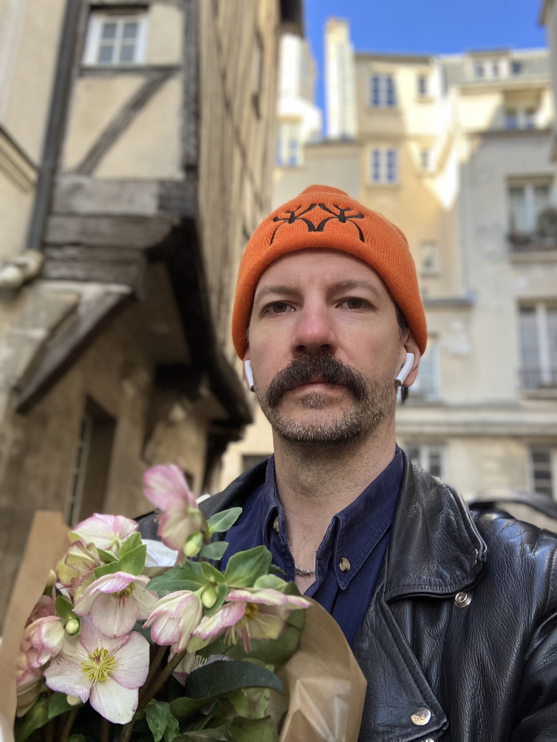 A close-up outdoor portrait of a person standing in a narrow historic street lined with old European buildings under a bright blue sky. They wear an orange knit beanie with a black graphic symbol, a dark leather jacket, and wireless earbuds, and hold a bouquet of pale pink flowers wrapped in brown paper.