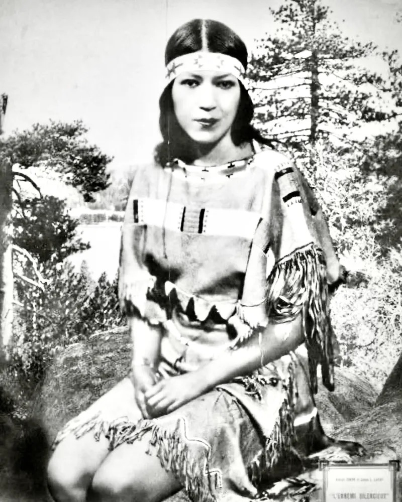 A vintage black-and-white portrait shows a young Indigenous woman seated outdoors in a wooded landscape. She wears a fringed garment with decorative geometric patterns and a beaded headband across her forehead. Her long dark hair is parted at the centre, and she faces the camera with a composed, direct expression.