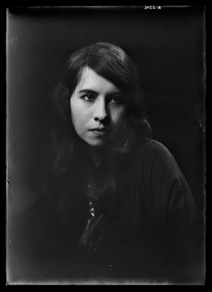 A moody black-and-white portrait of a young woman with long dark hair, photographed against a deep black background. Soft, directional lighting illuminates one side of her face, creating dramatic shadows and a contemplative atmosphere. Her direct gaze and neutral expression give the image an intimate, timeless quality reminiscent of early photographic portraiture or wet plate photography.