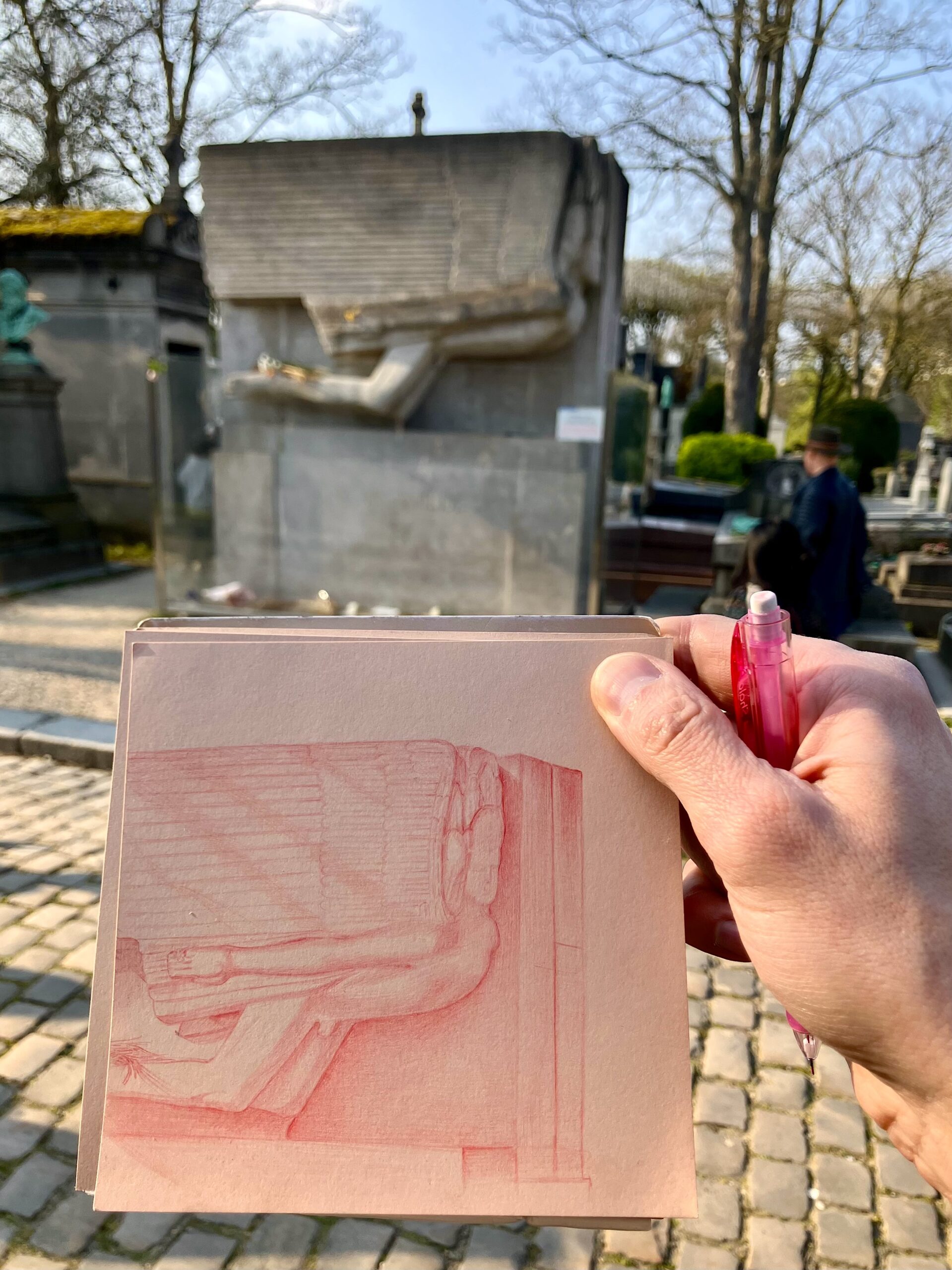 A hand holds a small sketchbook and a pink pen in the foreground of a sunlit cemetery scene. The sketch in the notebook appears to depict a reclining sculptural form, echoing the blurred stone monument in the background. Cobblestone paths, mausoleums, leafless trees, and a passerby in dark clothing create the atmosphere of a quiet historic cemetery, likely during early spring or late autumn.