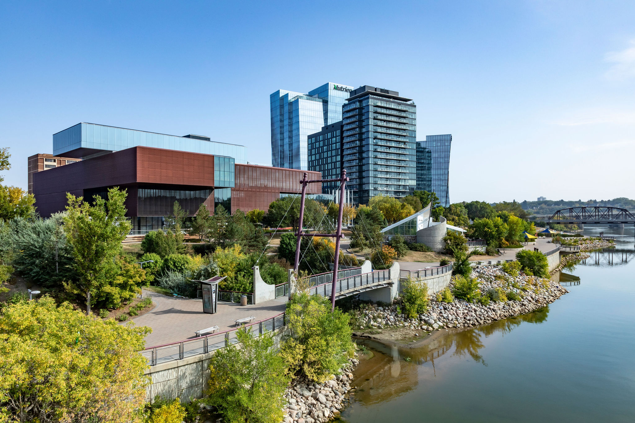 Exterior view of Remai Modern along the South Saskatchewan River in Saskatoon. The museum’s distinctive copper-toned architecture is framed by riverfront pathways, landscaped greenery, and nearby downtown towers under a clear blue sky.