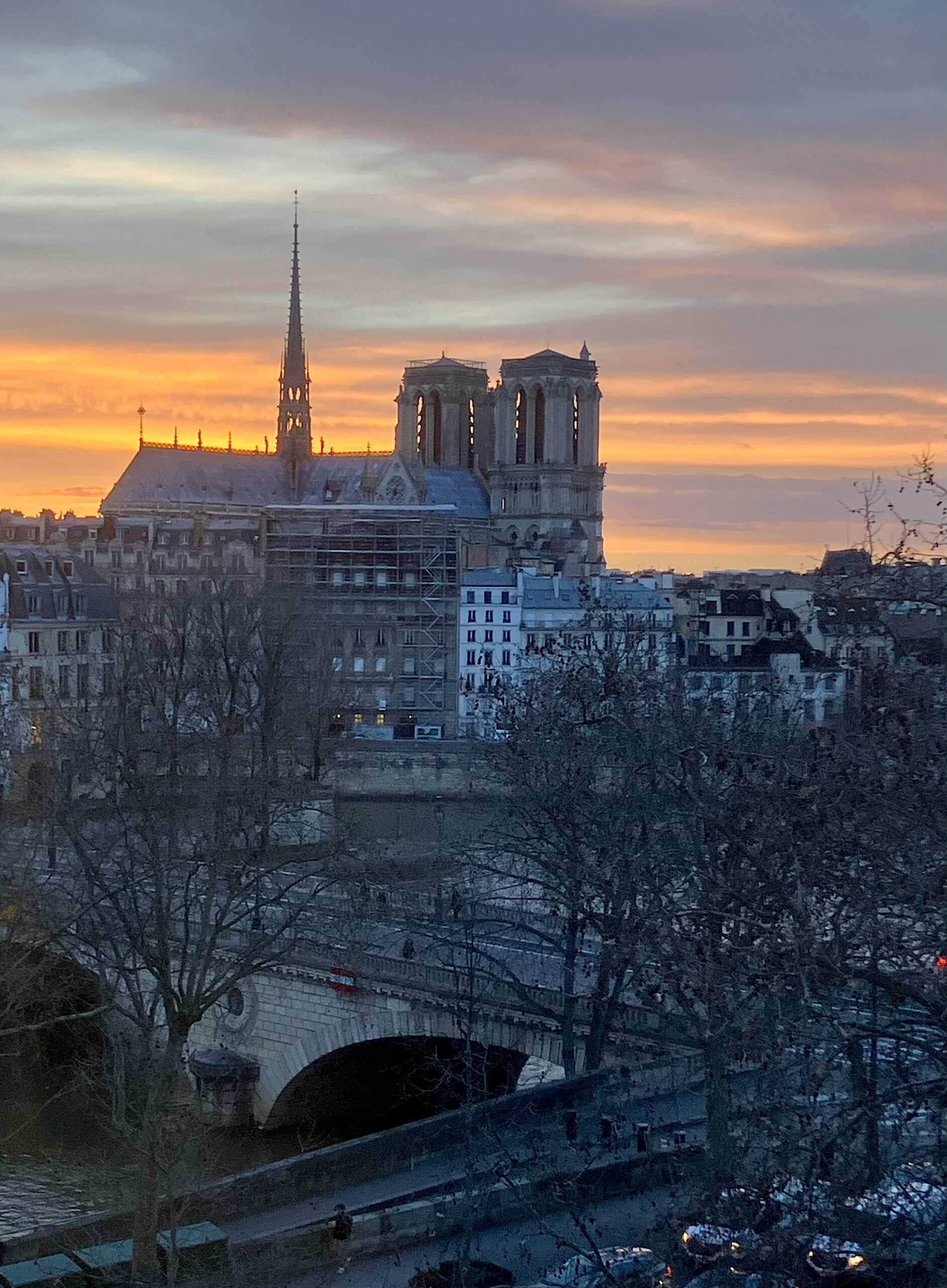 A sweeping sunset view over Paris featuring Notre-Dame Cathedral silhouetted against a glowing sky streaked with orange, pink, and blue clouds. In the foreground, bare winter trees and a stone bridge cross the Seine River, while historic buildings line the riverbanks below the cathedral’s towers and spire.