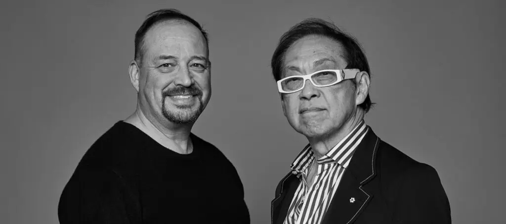 Portrait of architects Bruce Kuwabara and Alfred Waugh standing together in a black-and-white promotional photograph. Kuwabara smiles toward the camera while Waugh wears distinctive white glasses and a striped shirt beneath a dark blazer.