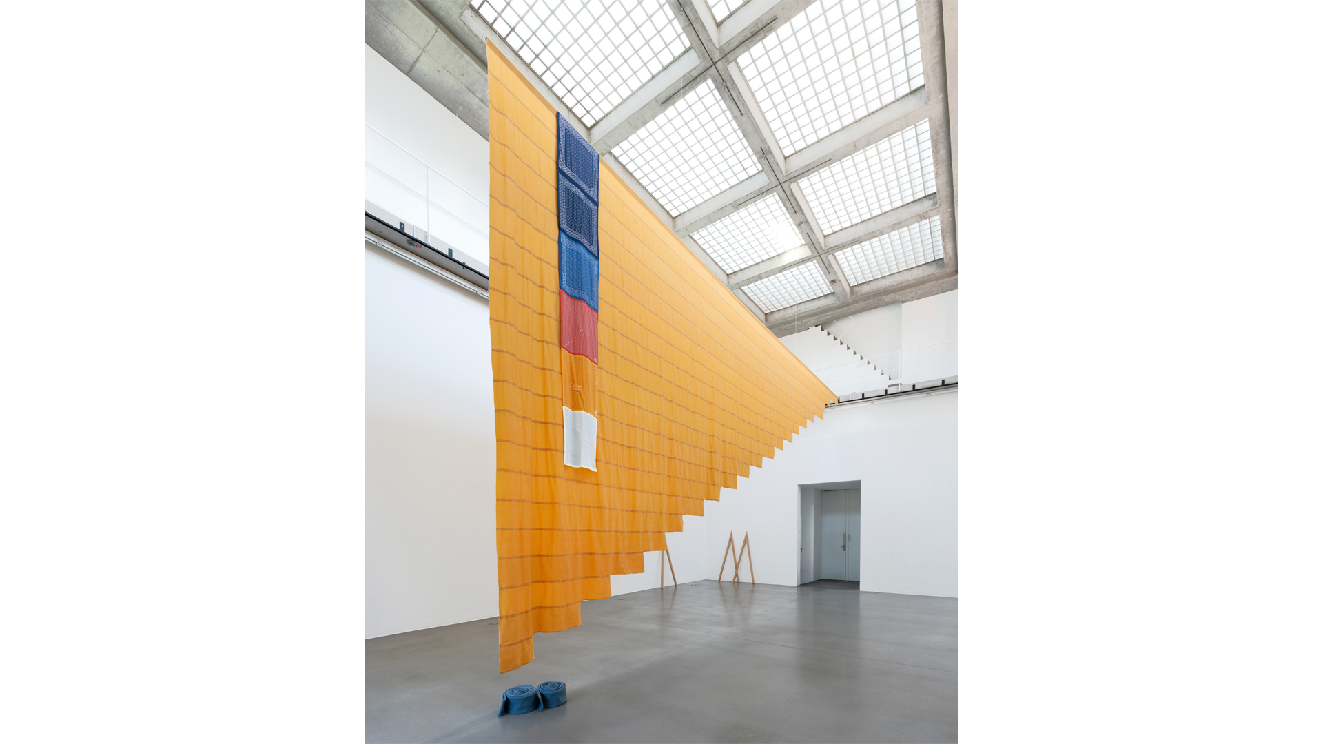A large orange textile installation hangs from the ceiling of a bright gallery, with a vertical strip of blue, red, orange and white fabric near one end.