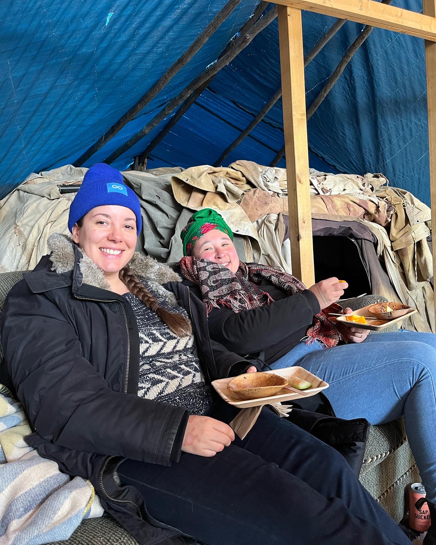 Two people in warm clothing smile while seated together with plates of food inside a rustic, tarp-covered shelter.
