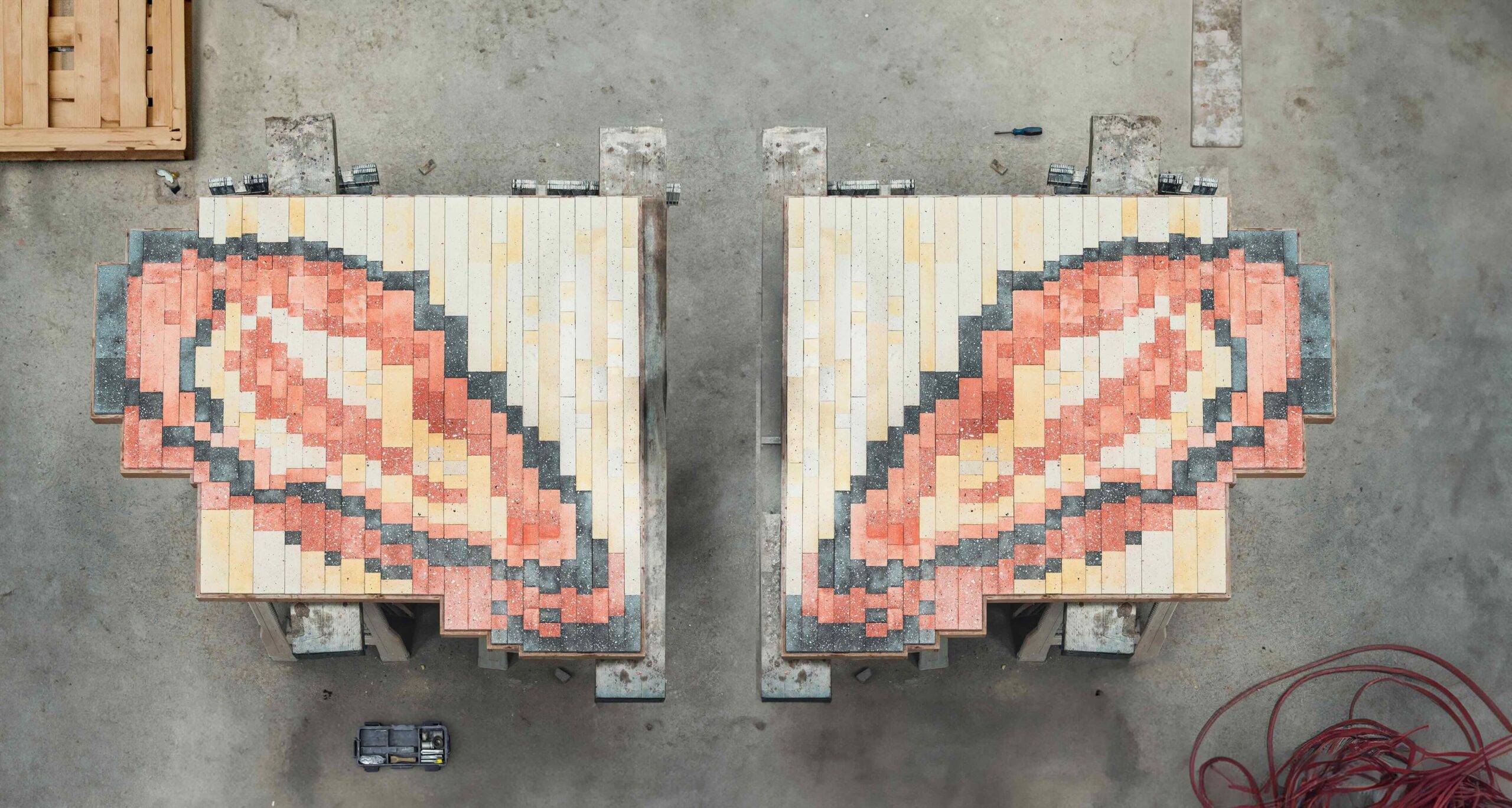 A photo of two tiled artworks that look like oyster shells in hues of grey, salmon and off white. They rest upon a grey concrete floor in this overhead shot that also features studio materials on the ground.