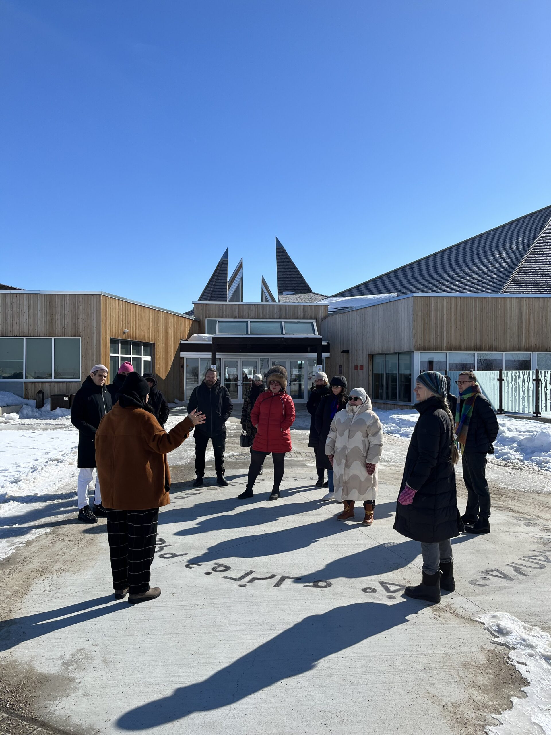 A group of people in winter clothing gather outdoors in a snowy courtyard, listening to a speaker in front of a large wood-clad building.