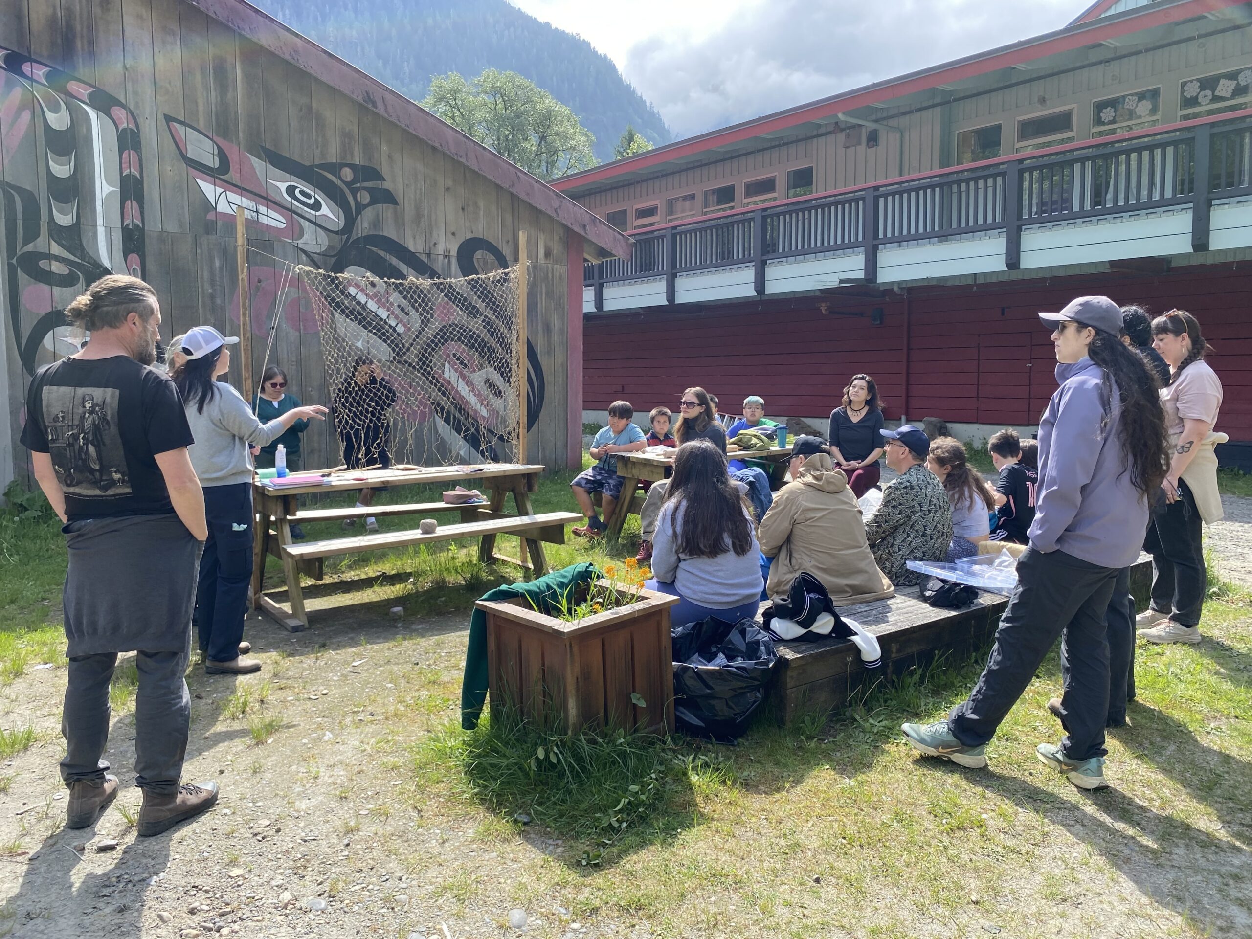 A group of adults and children gather outdoors for a oolican net demonstration beside a wooden building painted with a large Indigenous-style mural, with mountains rising in the background.