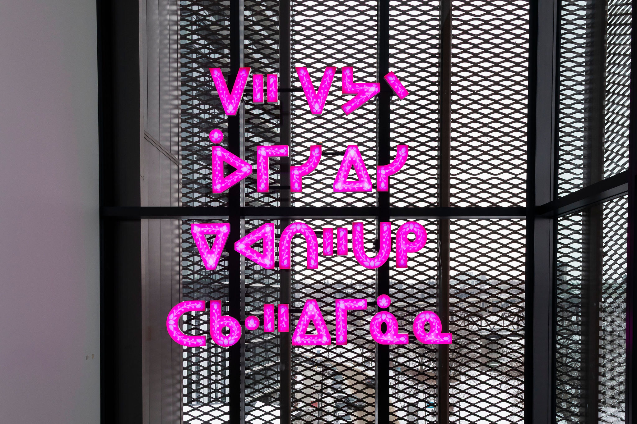 A bright pink LED artwork featuring stylized syllabic characters glows against a large window covered by a dark metal lattice.