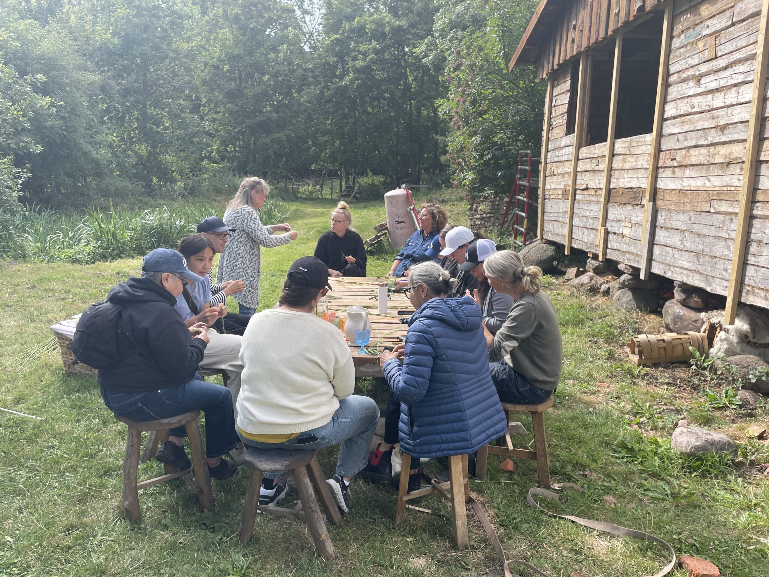 A group of people sit around a long wooden table outdoors, participating in a stinging nettle weaving workshop beside a rustic wooden building surrounded by trees and greenery.