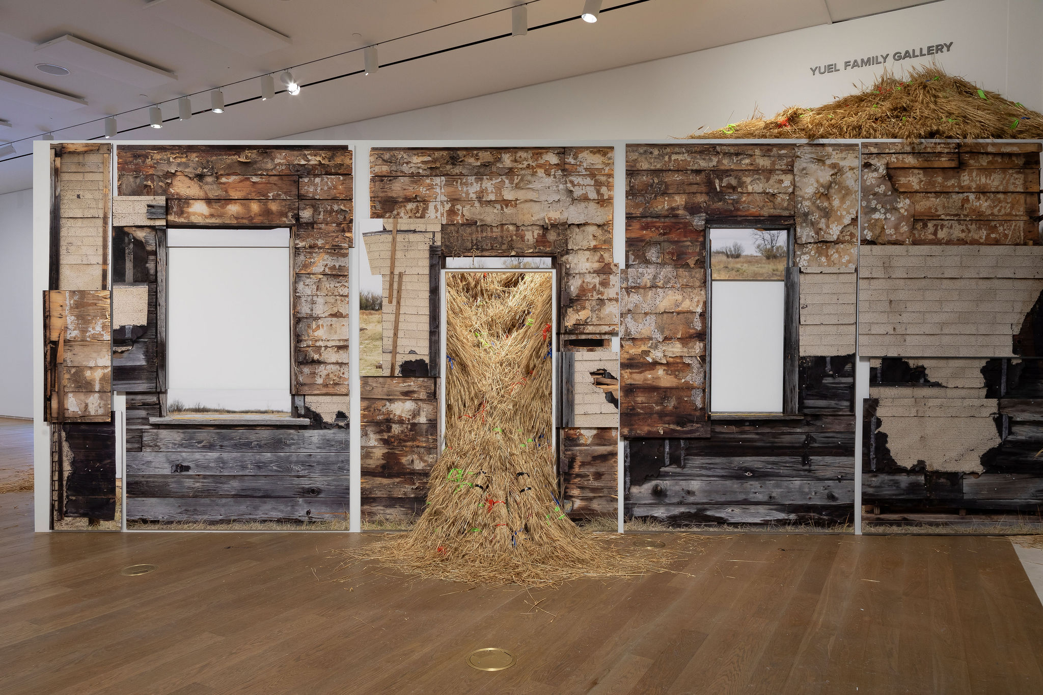 A gallery installation recreates the weathered walls of a wooden building, with openings filled by images of a rural landscape and a large mound of hay spilling through the centre doorway.