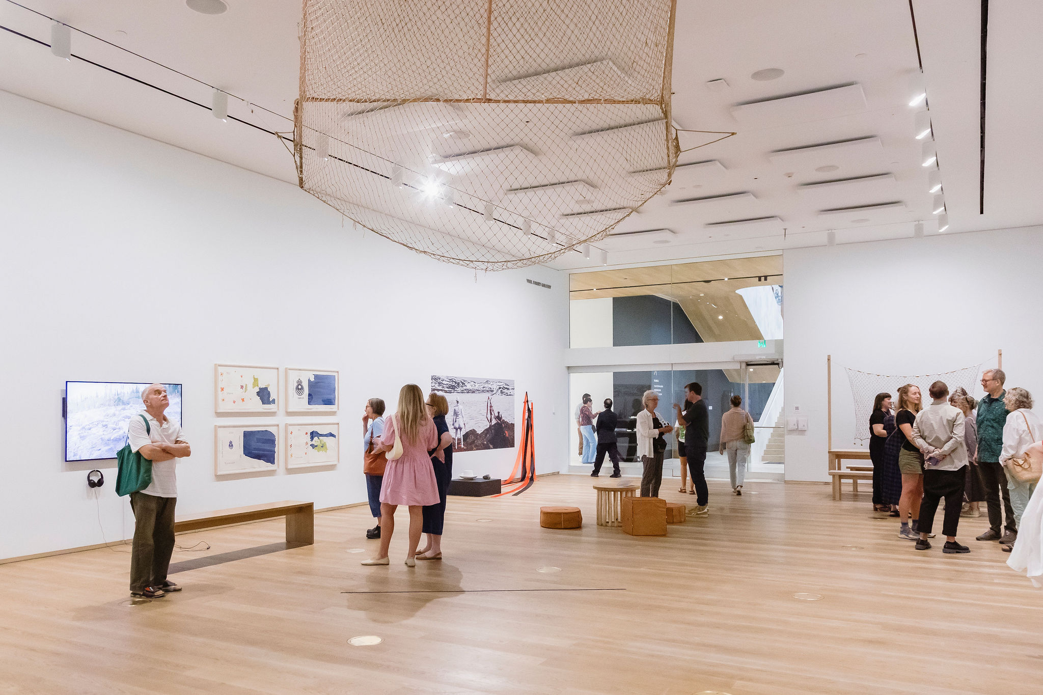 Visitors gather and converse in a spacious gallery displaying photographs, drawings, video and suspended net-like installations.