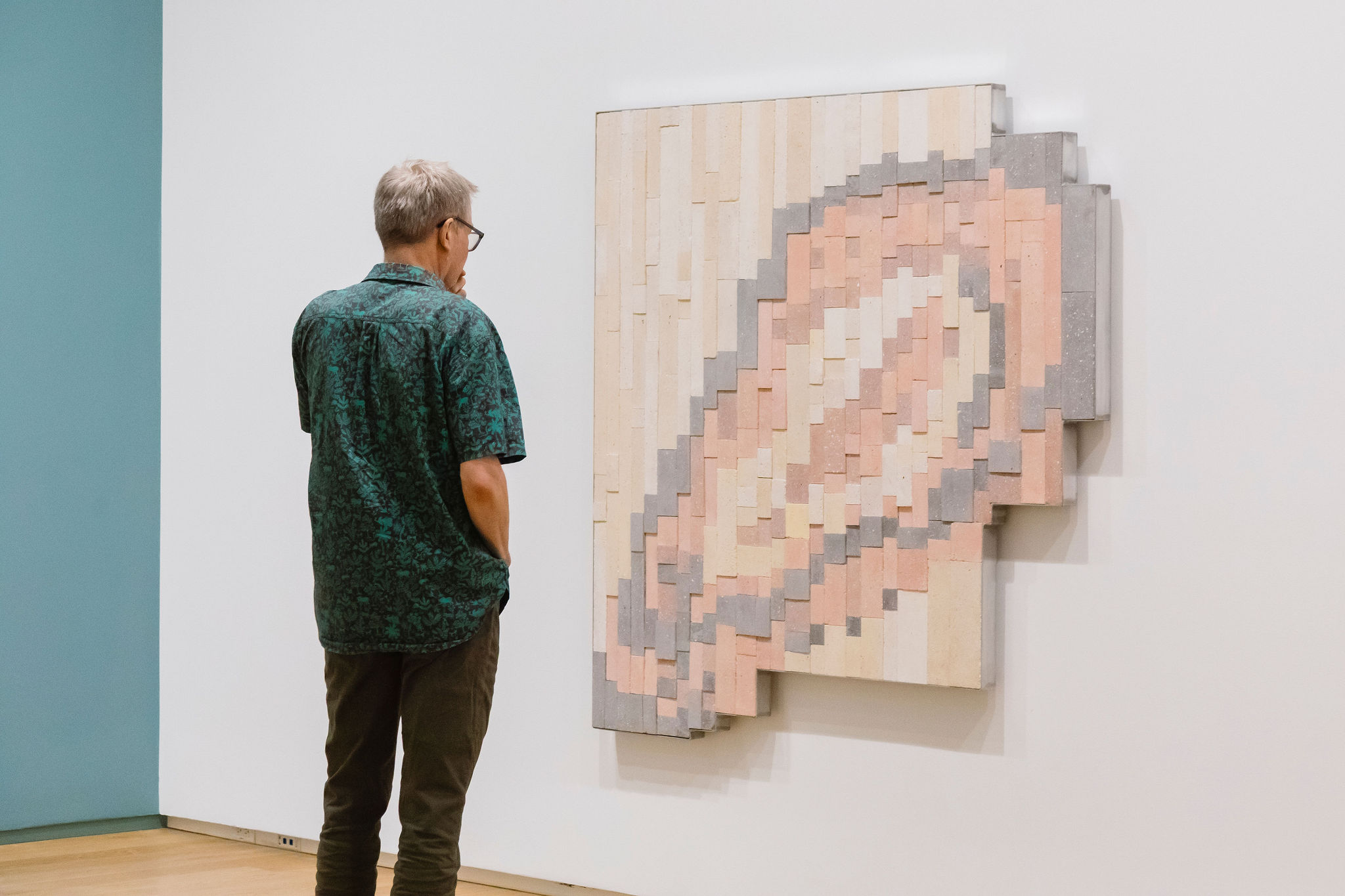 A visitor stands in a gallery, looking closely at a large, textured geometric artwork composed of overlapping blocks in pale pink, cream and grey.