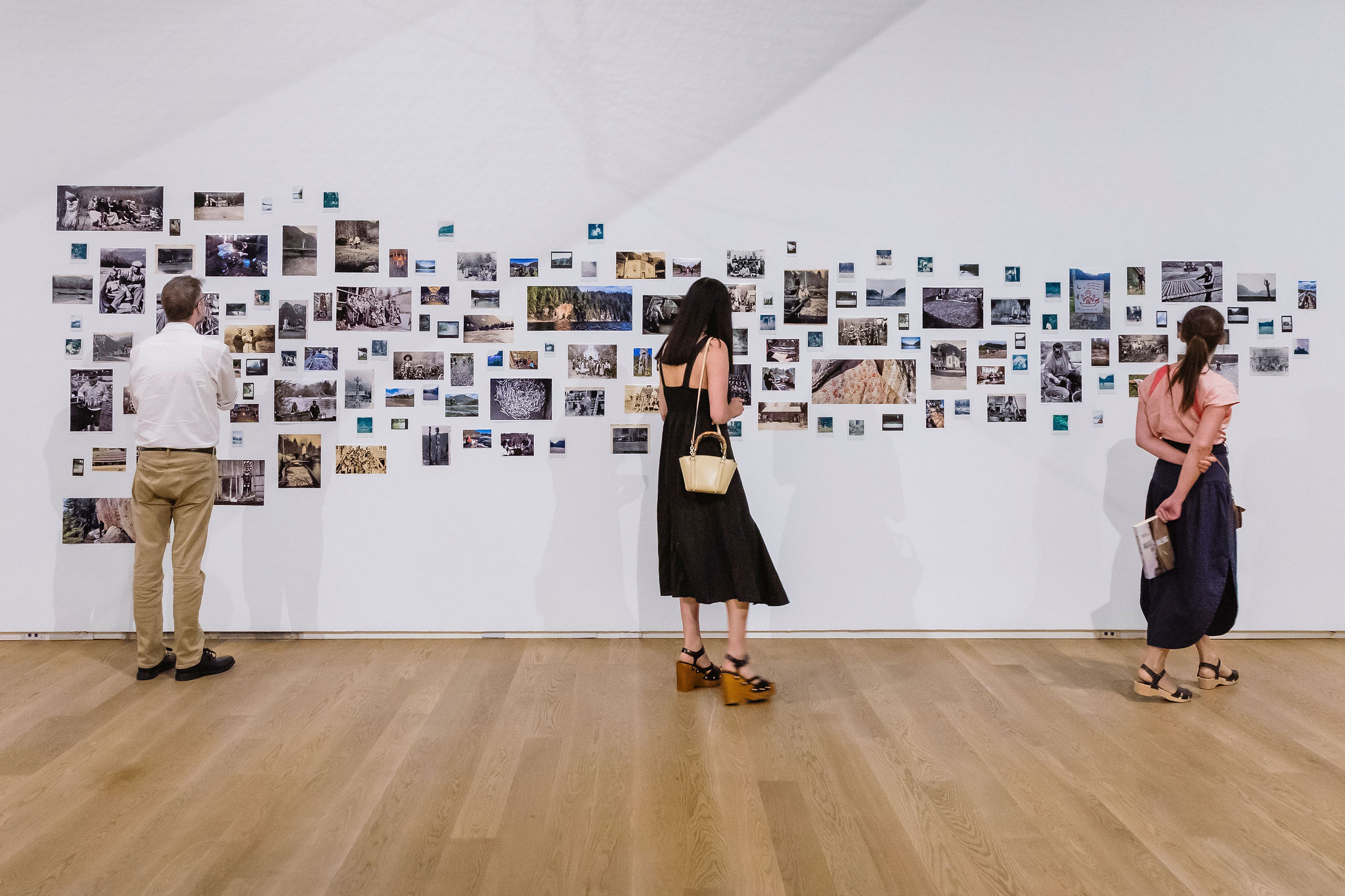 Three visitors view a large installation of photographs arranged across a white gallery wall.