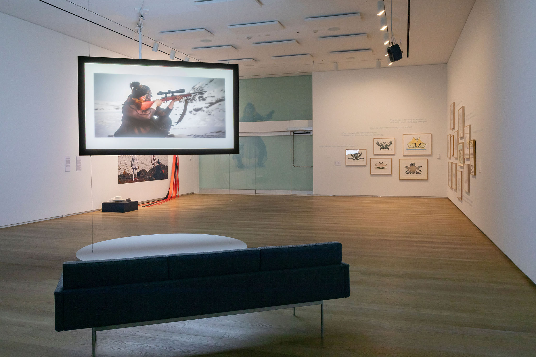 A gallery installation features a suspended illuminated image of a person aiming a rifle in a snowy landscape, with framed artworks displayed on the surrounding walls.