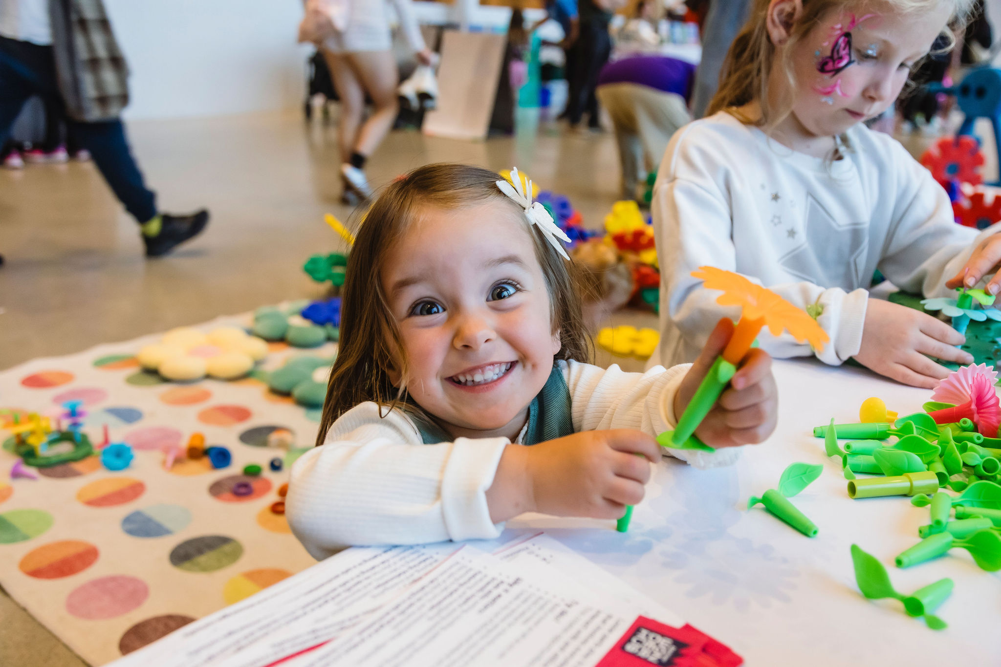 A young child grins at the camera while building with green and orange plastic flower pieces at a craft table.