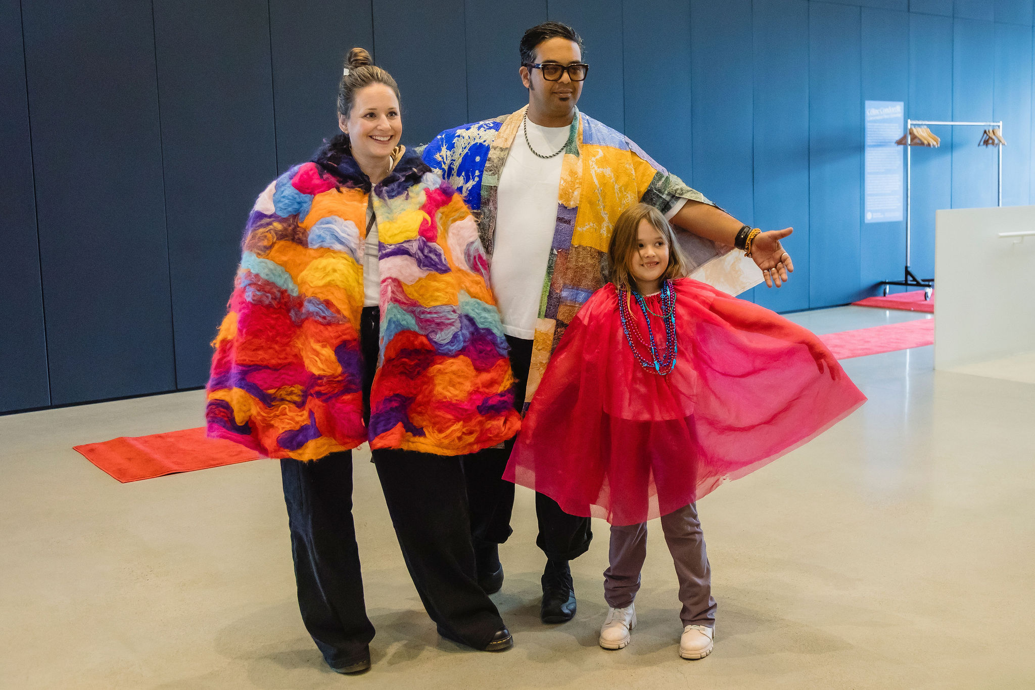 Two adults and a child pose in colourful handmade capes on a red runway in a blue-walled gallery.
