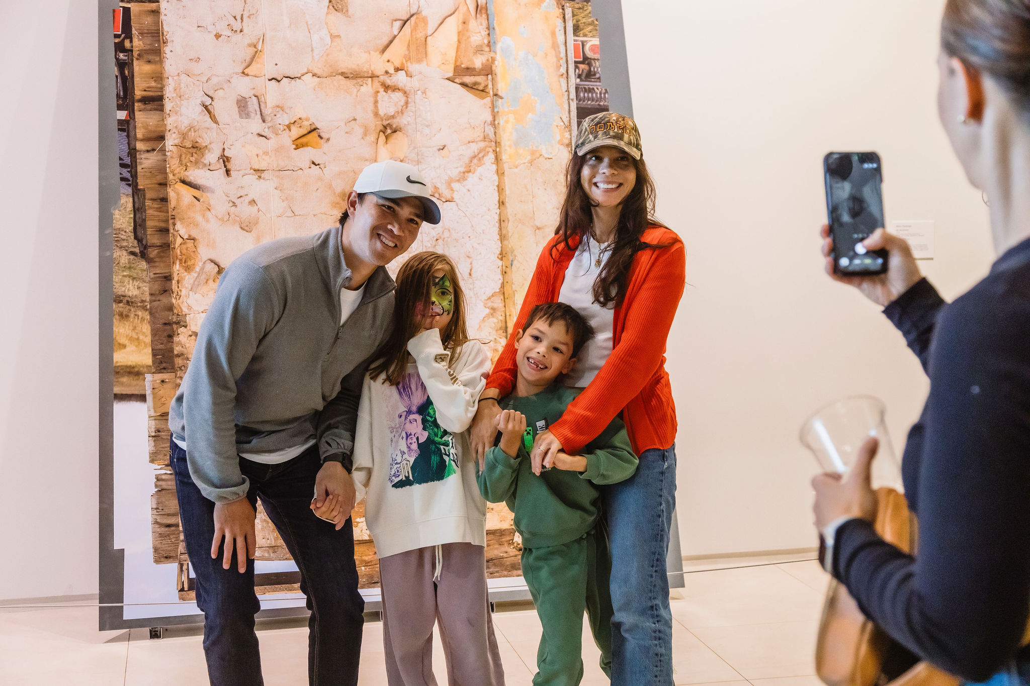A family of four smiles for a photo in front of a large artwork while someone takes their picture on a phone.