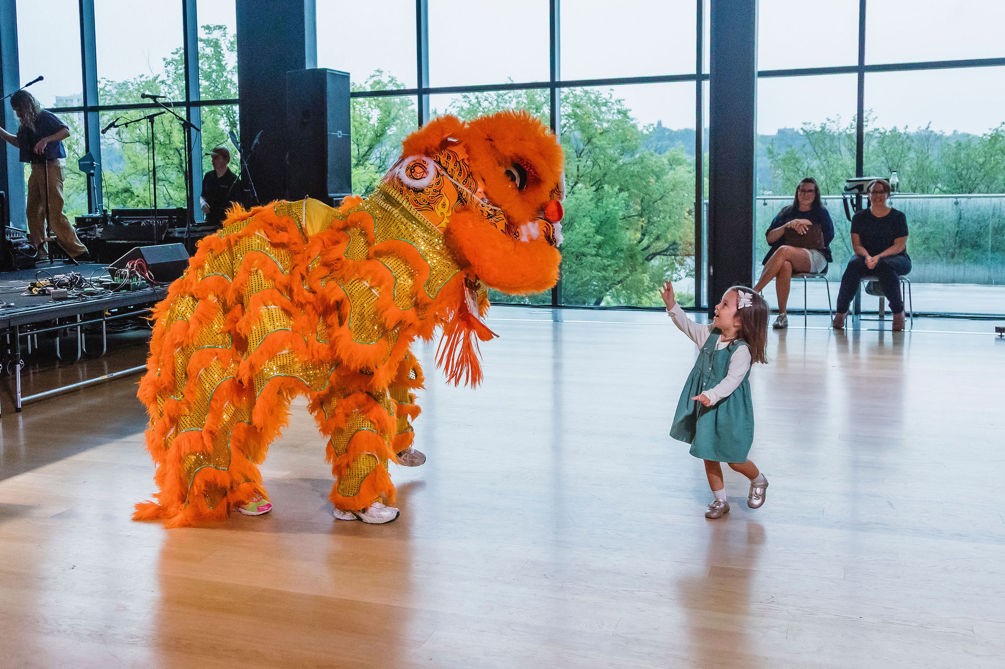A small child reaches up toward a large orange lion dance costume in a bright, window-lined gallery.