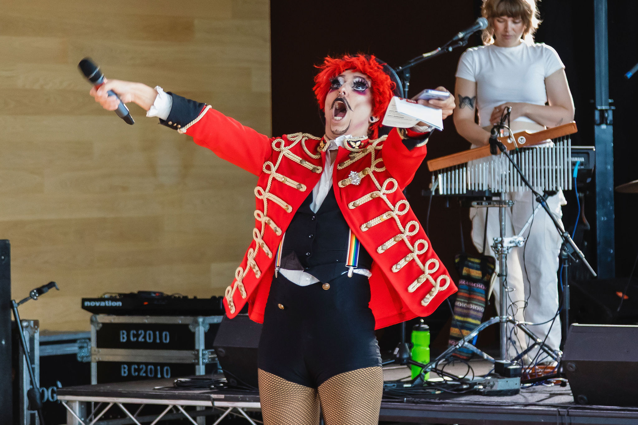 A performer in a red ringmaster jacket and red wig throws an arm out mid-announcement, microphone in hand.