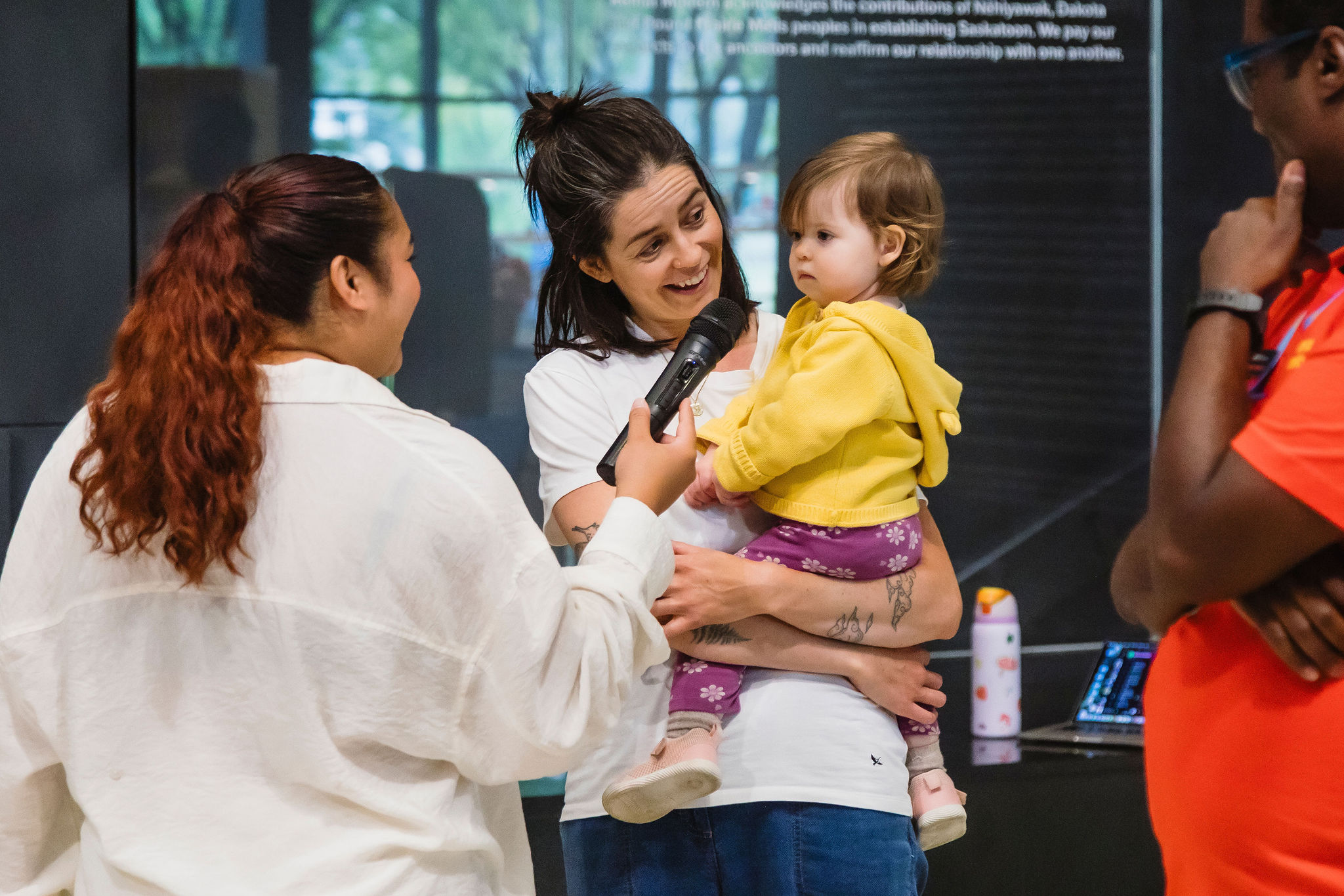 A person holding a toddler on one hip speaks into a microphone, smiling at someone beside them.