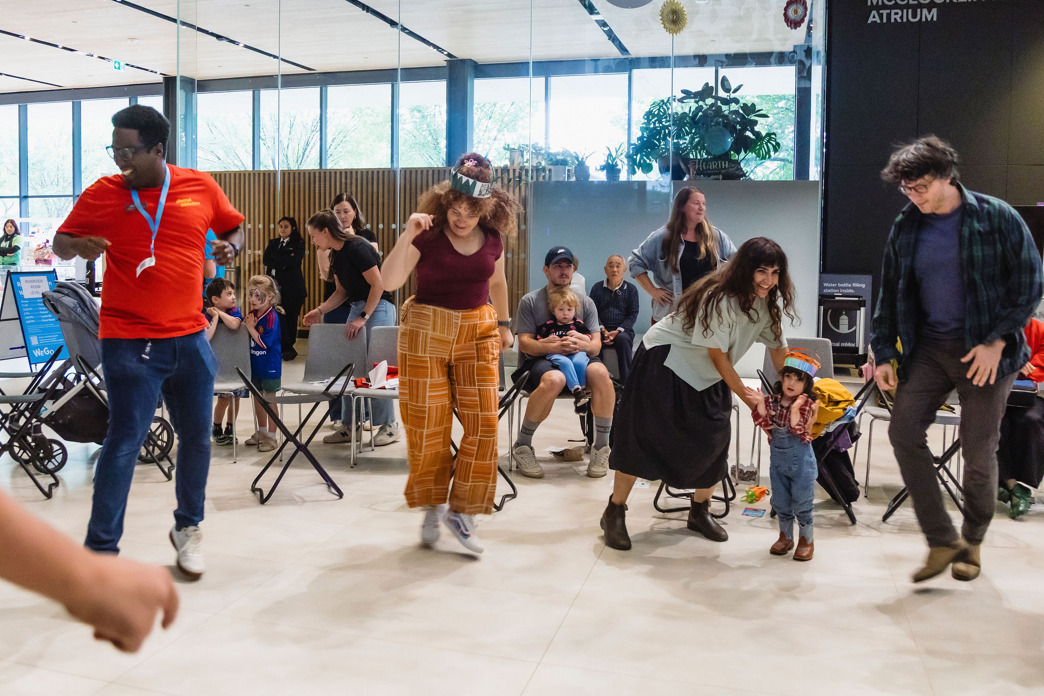 Four adults and a young child dance together in an open area of Remai Modern's atrium while seated visitors watch from a circle of chairs.