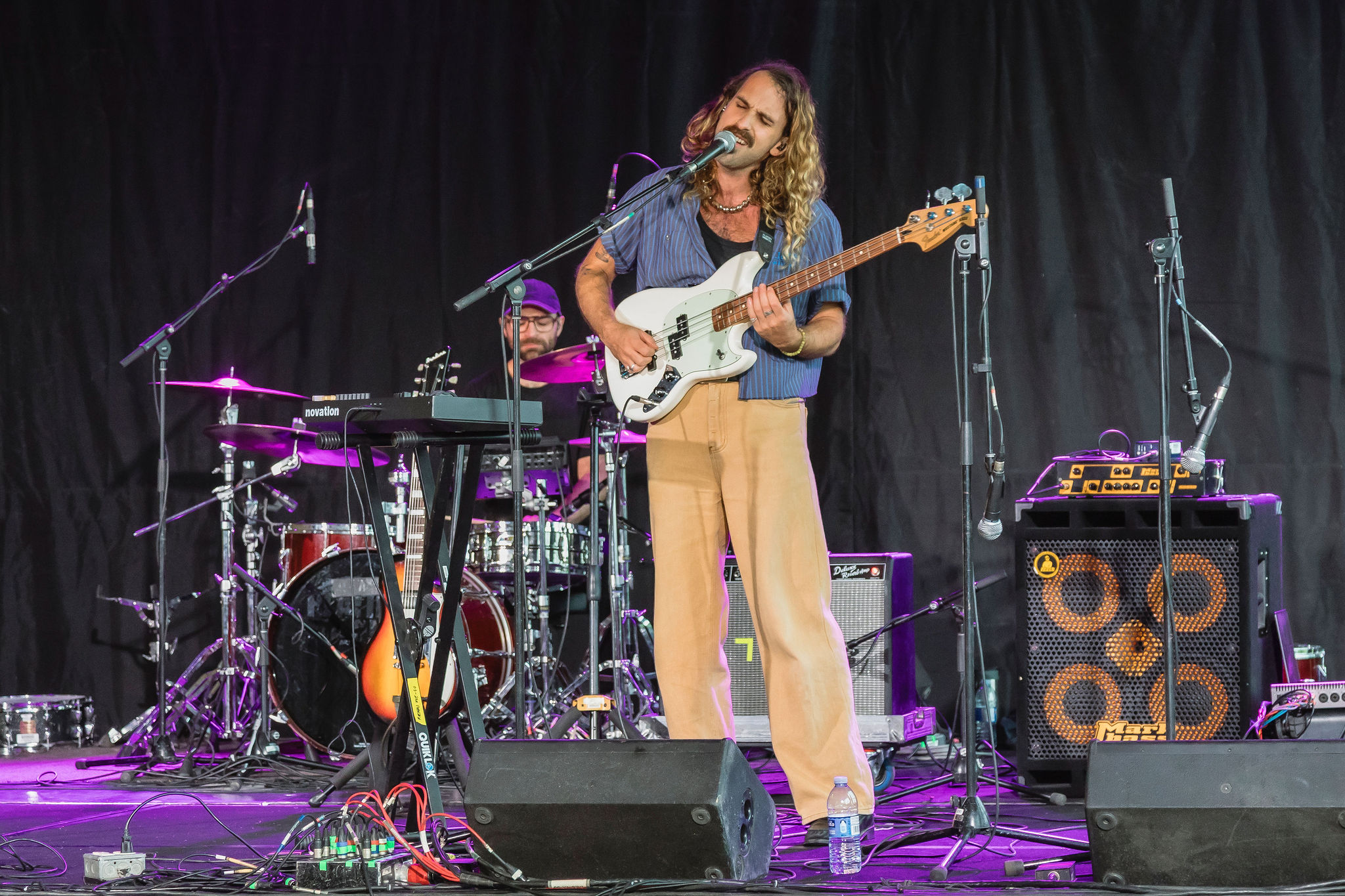 A bassist sings on a purple-lit stage with a drummer behind him.